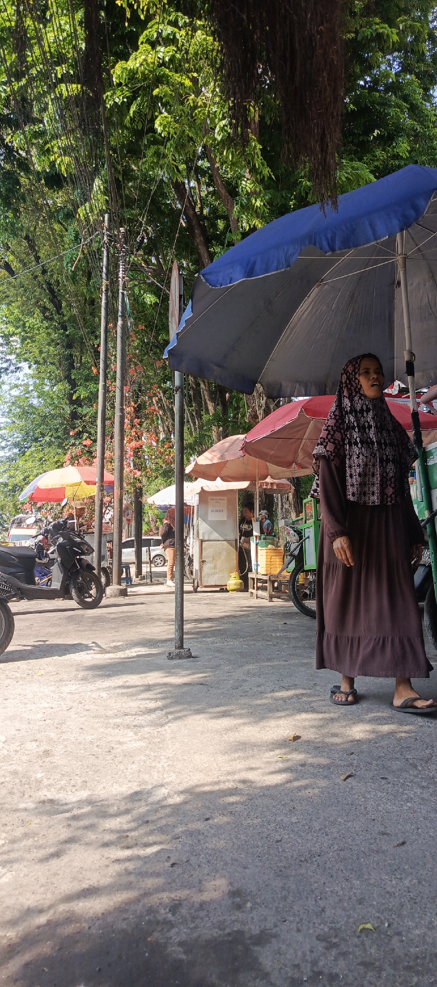 The image depicts a street scene in Krian, Indonesia, likely on a sunny day with ample shade provided by large trees and umbrellas. In the foreground, a woman wearing a dark brown dress and a patterned headscarf is walking on a paved surface. She is under the shade of a large blue umbrella and a smaller dark gray umbrella. To her left, several colorful umbrellas are visible, covering market stalls. In the middle ground, motorcycles are parked, and there are various food carts or stalls. One stall has a sign that reads "TERANG BULAN" and "JASUKE". A person is visible behind this stall, possibly serving customers. To the right of the central stall, a green cart with yellow containers is parked. The background features lush green trees with hanging vines, suggesting a tropical environment. Power lines and poles are visible among the trees. The overall impression is of a bustling outdoor market or roadside food vendors operating in a shaded area.
