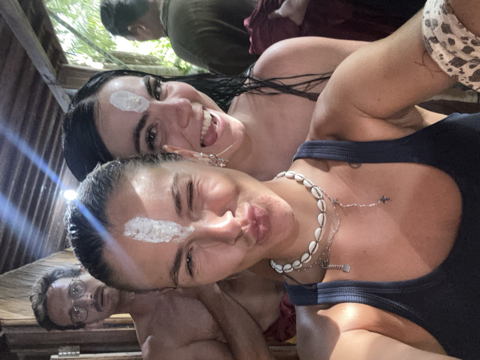 The image captures four individuals inside a wooden structure, likely in Punggul, Indonesia. In the immediate foreground, a light-skinned female with wet, dark hair and a vertical white marking on her forehead winks and forms a "kiss" expression. She wears a dark blue tank top, a cowrie shell choker, and layered silver necklaces. Behind her to the right, a second light-skinned female with wet, dark hair and an identical white forehead marking smiles broadly. Her left ear displays multiple small hoop earrings. In the left background, a male figure with glasses and a mustache, shirtless and wearing a dark red sarong-like garment, looks towards the camera with a neutral expression. A fourth male figure is partially visible in the right background, facing away from the camera, wearing a dark shirt and a red garment. The environment features exposed wooden beams and planks, with bright natural light entering from above and lush green foliage visible through an opening in the background right.