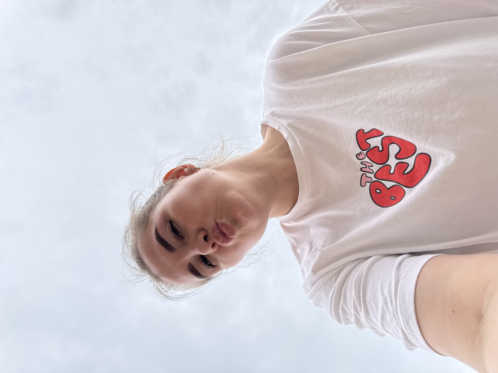 A woman with light skin and blonde hair is angled downwards with a light-colored sky as the backdrop. She is wearing a white t-shirt with the words "THE BEST" in red lettering on the upper right side. Her lips are pursed. The photograph seems to be taken at a low angle, looking upwards towards the subject. No specific indicators suggest this photo was taken in Gornyi (Dvurechenskoe S/P), Russia.