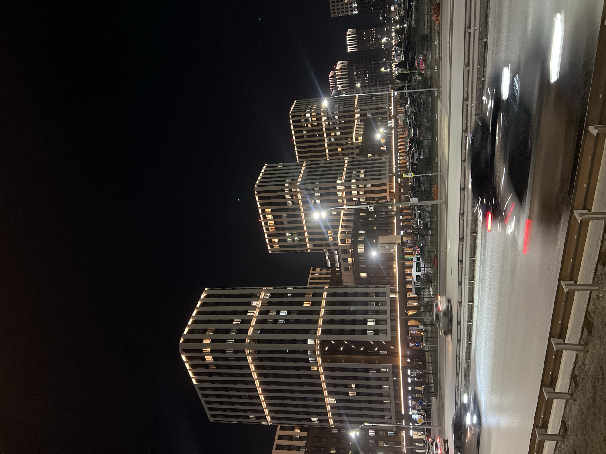 The image depicts a nighttime urban scene in Kazan, Russia, showing modern residential buildings illuminated with horizontal stripes of light. A road runs parallel to the buildings, with blurred car headlights and taillights indicating vehicle movement. Streetlights are visible along the road and parking area with cars. The sky is dark and clear.