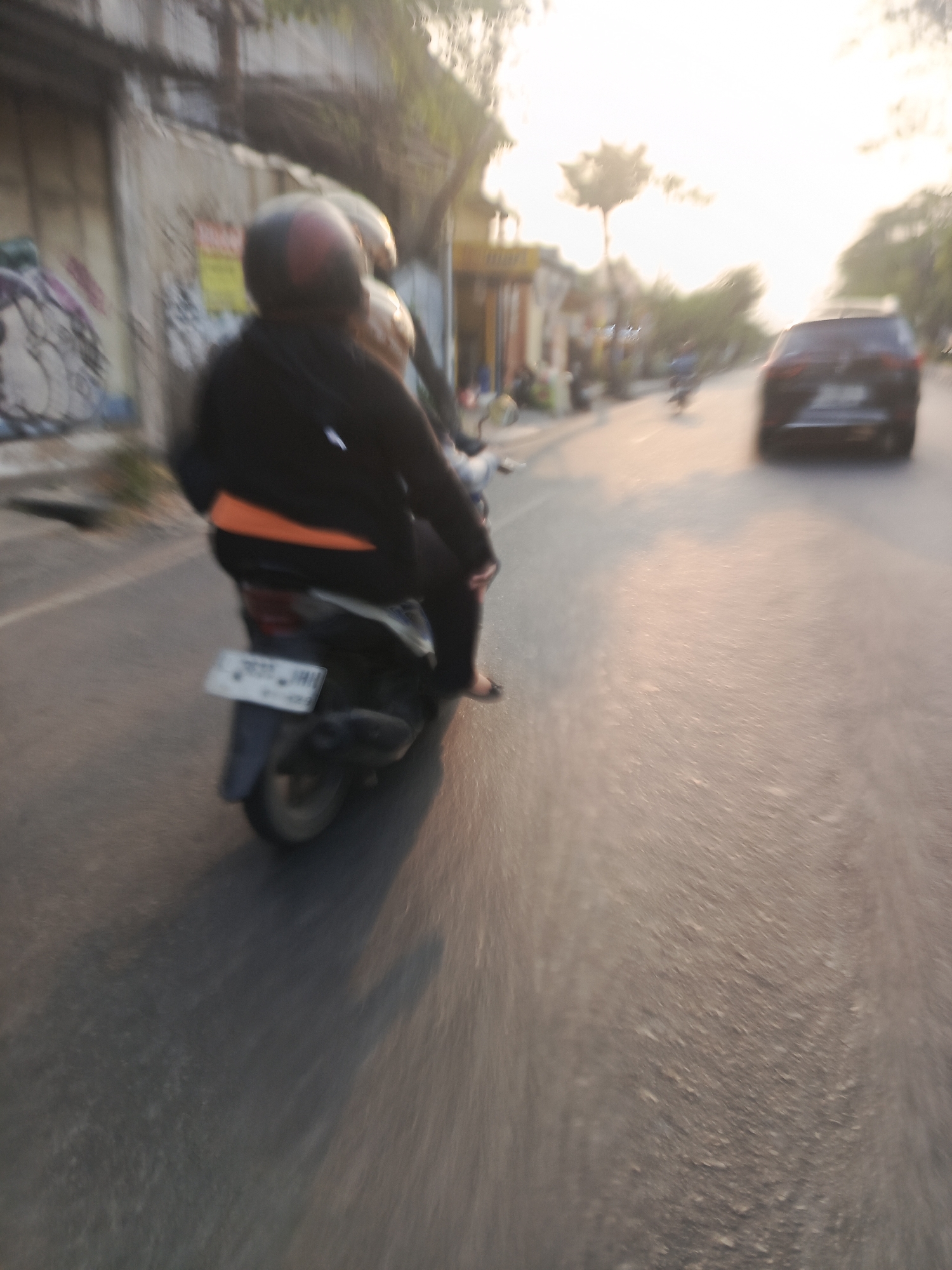 The image captures a motion-blurred view of a street scene in Surabaya, Indonesia, likely during the late afternoon or early evening, as indicated by the warm, low-angle sunlight casting long shadows. In the foreground, a person wearing a black jacket and a helmet is riding a scooter. The scooter has a visible license plate with the characters "BE 7637 JMH". The rider is wearing an orange garment visible beneath the jacket. To the right of the scooter, a dark-colored SUV is also in motion. The street is paved and appears to be in moderate condition. On the left side of the road, there are buildings with graffiti on their walls. The background shows more buildings, trees, and other vehicles, suggesting a typical urban environment. The overall impression is one of movement and daily life in a city.