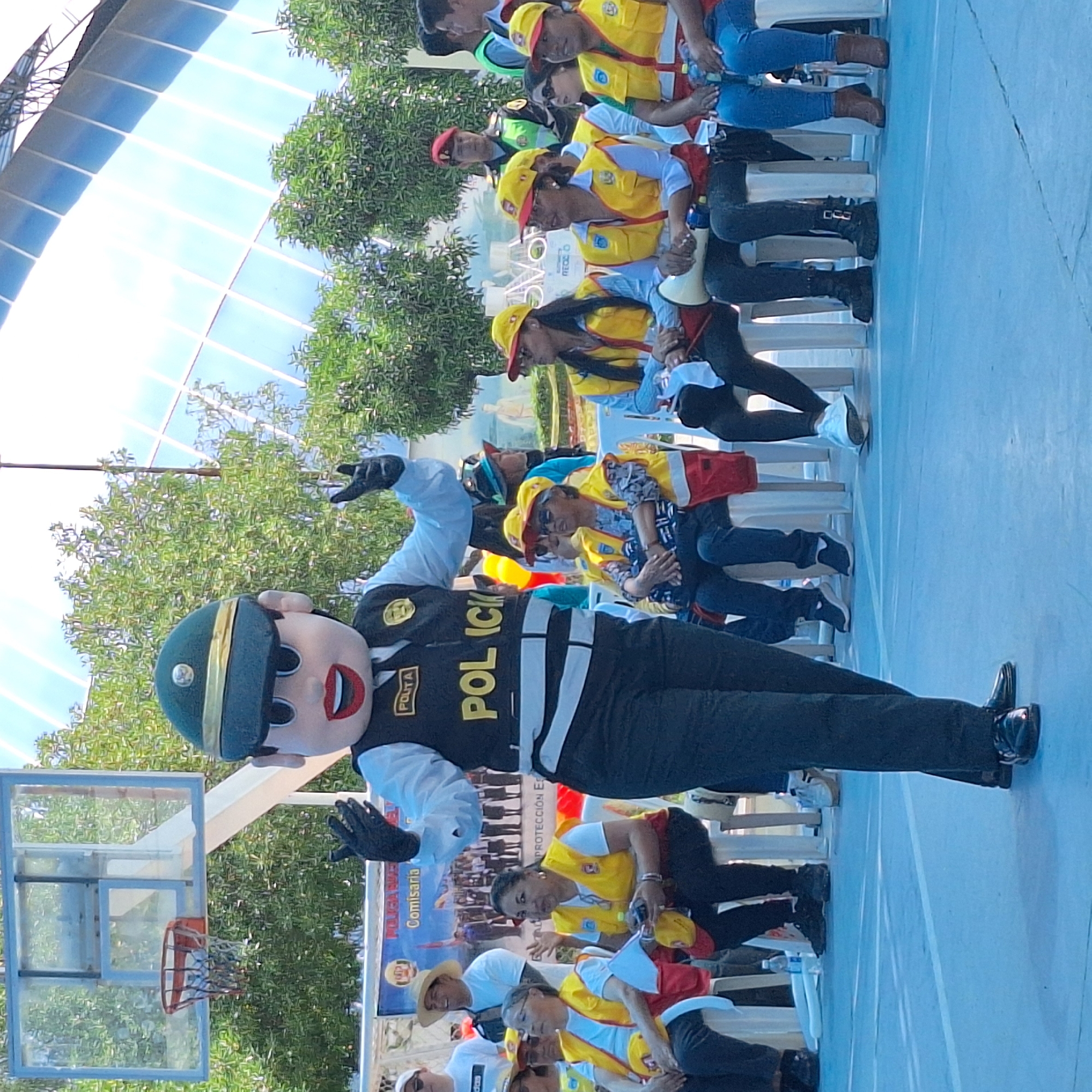 The image depicts a medium shot of a mascot dressed as a police officer, with the word "POLICIA" displayed prominently on its chest. The mascot is standing on a blue surface with white lines, possibly a basketball court. Behind the mascot, there is a basketball hoop with a net.
In the background, there are several people sitting in chairs. Many of them are wearing yellow vests and caps. A banner is visible behind the seated people, though the text is partially obscured.
The location appears to be outdoors, under a semi-transparent structure that lets in sunlight. Trees are visible behind the structure.
The city where the photograph was taken is José Luis Bustamante y Rivero, Peru.