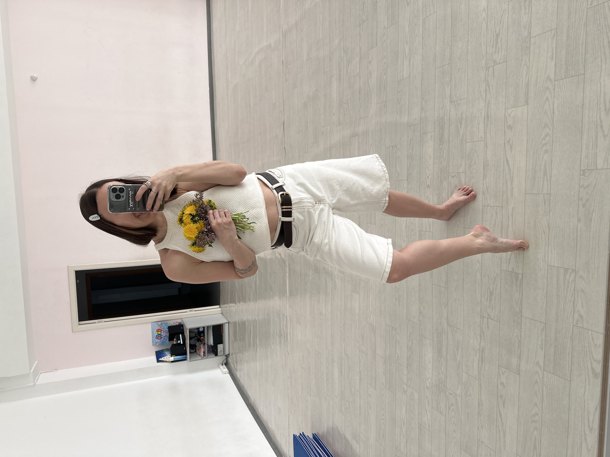 A person is reflected in a mirror, holding a bouquet of flowers and a smartphone. The person is wearing a white knit top, white denim shorts with a dark brown belt, and a silver hair clip. The room contains a door with a black frame, white walls, and white flooring. Below the door is a white storage unit containing various items. The location is Votkinsk, Russia.