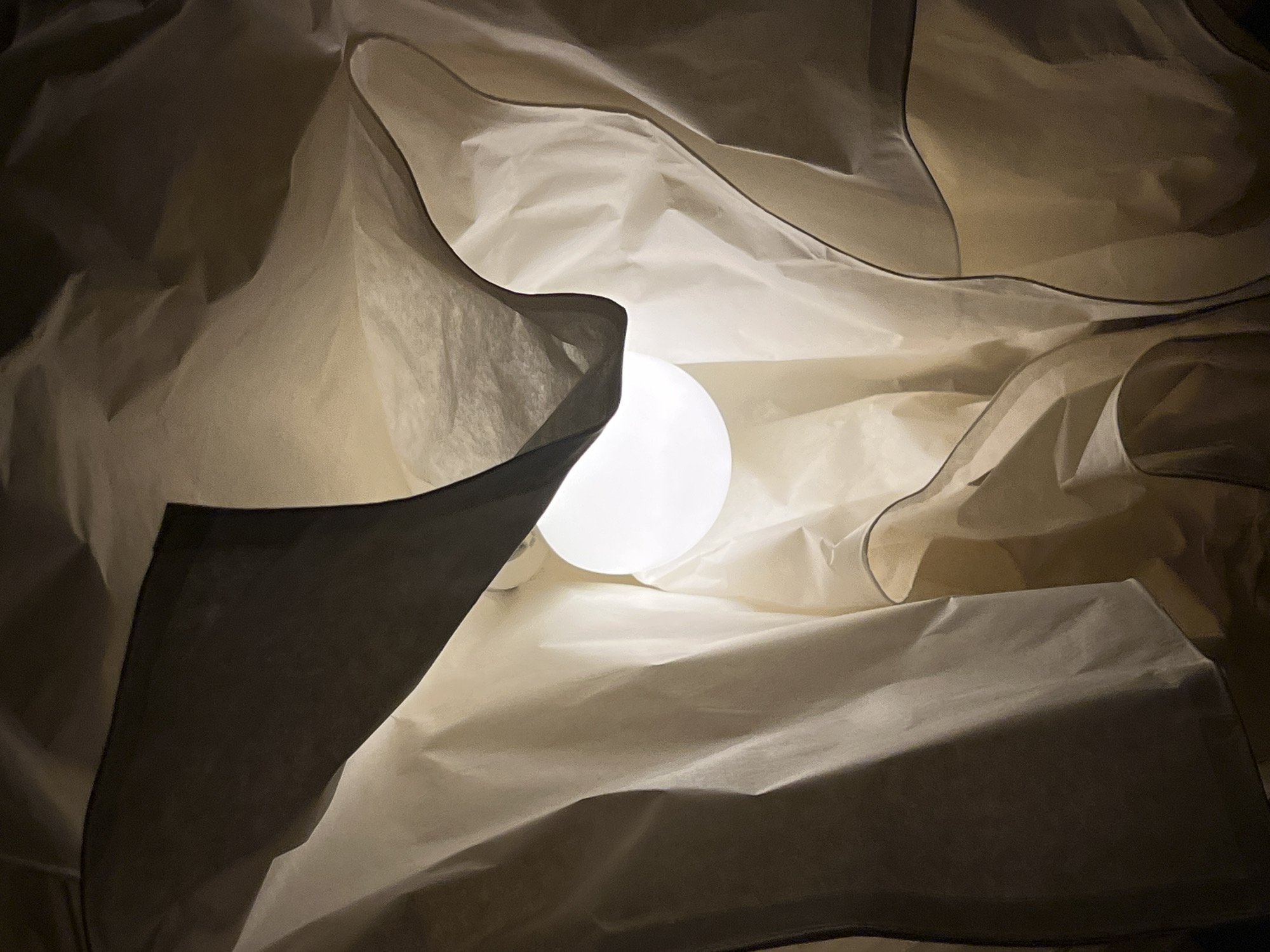 The image presents an abstract, close-up view of a light fixture, likely a paper or fabric lantern, seen from an inward perspective. The central subject is a brightly glowing, spherical white light bulb, emitting a soft, warm light. Surrounding the bulb are multiple layers of translucent, off-white material, resembling crumpled or folded paper or thin fabric. These layers create dynamic patterns of light and shadow, with lighter areas directly illuminated by the bulb and darker, muted beige and brown tones in areas that are folded over or further from the light source. The edges of some of the material folds are outlined by subtle darker lines, enhancing the sense of depth and form.
There are no people or discernible activities taking place. The setting is ambiguous but suggests an indoor, intimate space, possibly a home or an art installation, designed to evoke a calm or artistic atmosphere. The lighting indicates it is likely nighttime or an interior space where artificial light is the primary source. No specific time of day, weather, or location cues are visible within the image itself. There is no visible text. The overall impression is one of soft illumination and textural intrigue.