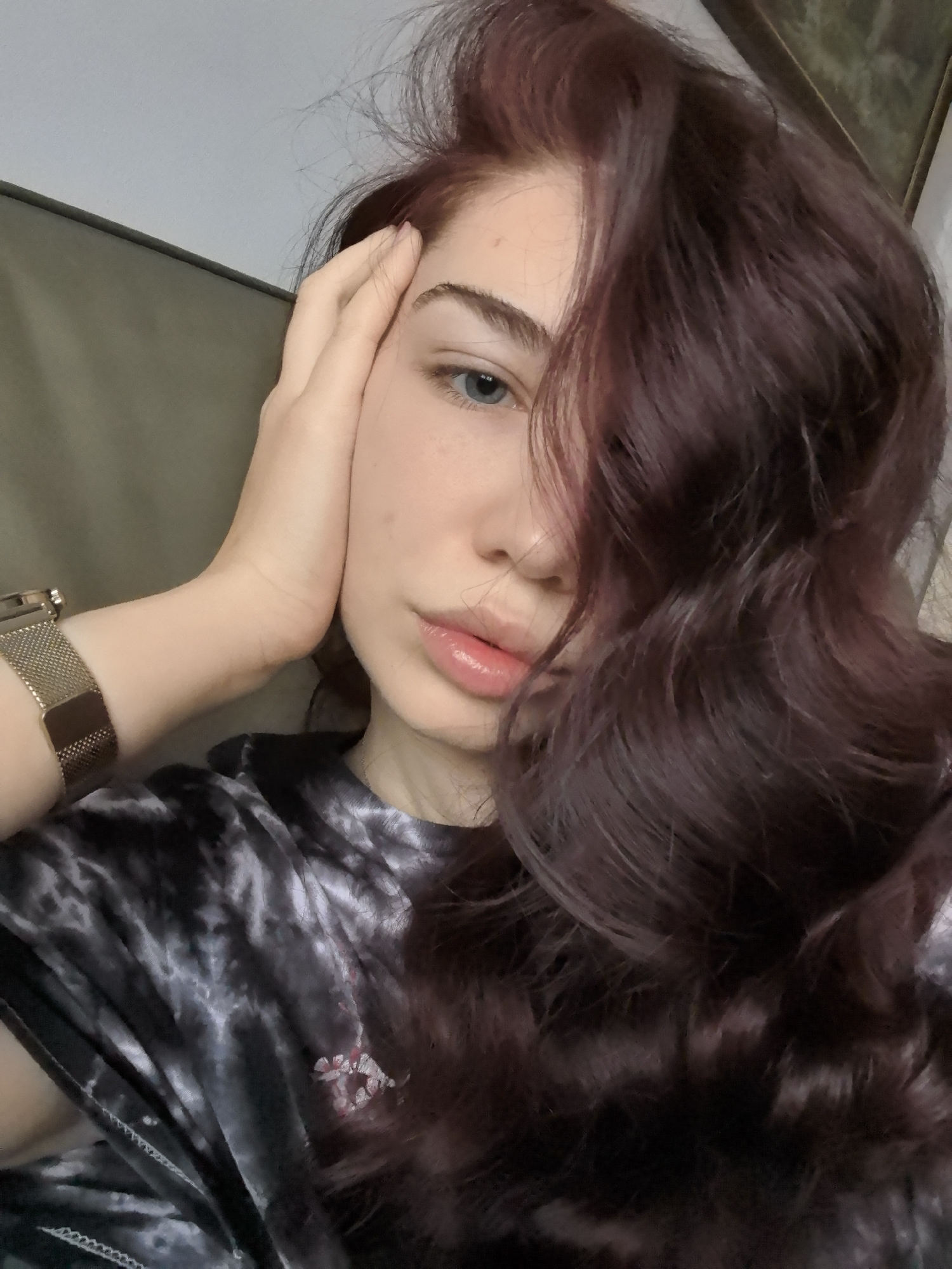 A close-up indoor portrait features a female subject. Her long, wavy, dark reddish-brown hair is styled to fall across the right side of her face. Her visible left eye is blue, and her lips are full and glossy pink. Her complexion is fair with visible freckles. The subject's right hand, with unpolished nails, is raised and positioned with fingers splayed against her right temple and cheek. She is wearing a dark grey and white tie-dye patterned top with a small, indistinct white and pink graphic on the chest. A gold-colored mesh strap watch is present on her left wrist. The background is indistinct, showing a light grey wall in the upper left, an olive-green upholstered surface below it, and a partially visible dark-framed object in the upper right. The specific location within the city of Stara Zagora, Bulgaria, cannot be determined from the visual information presented.