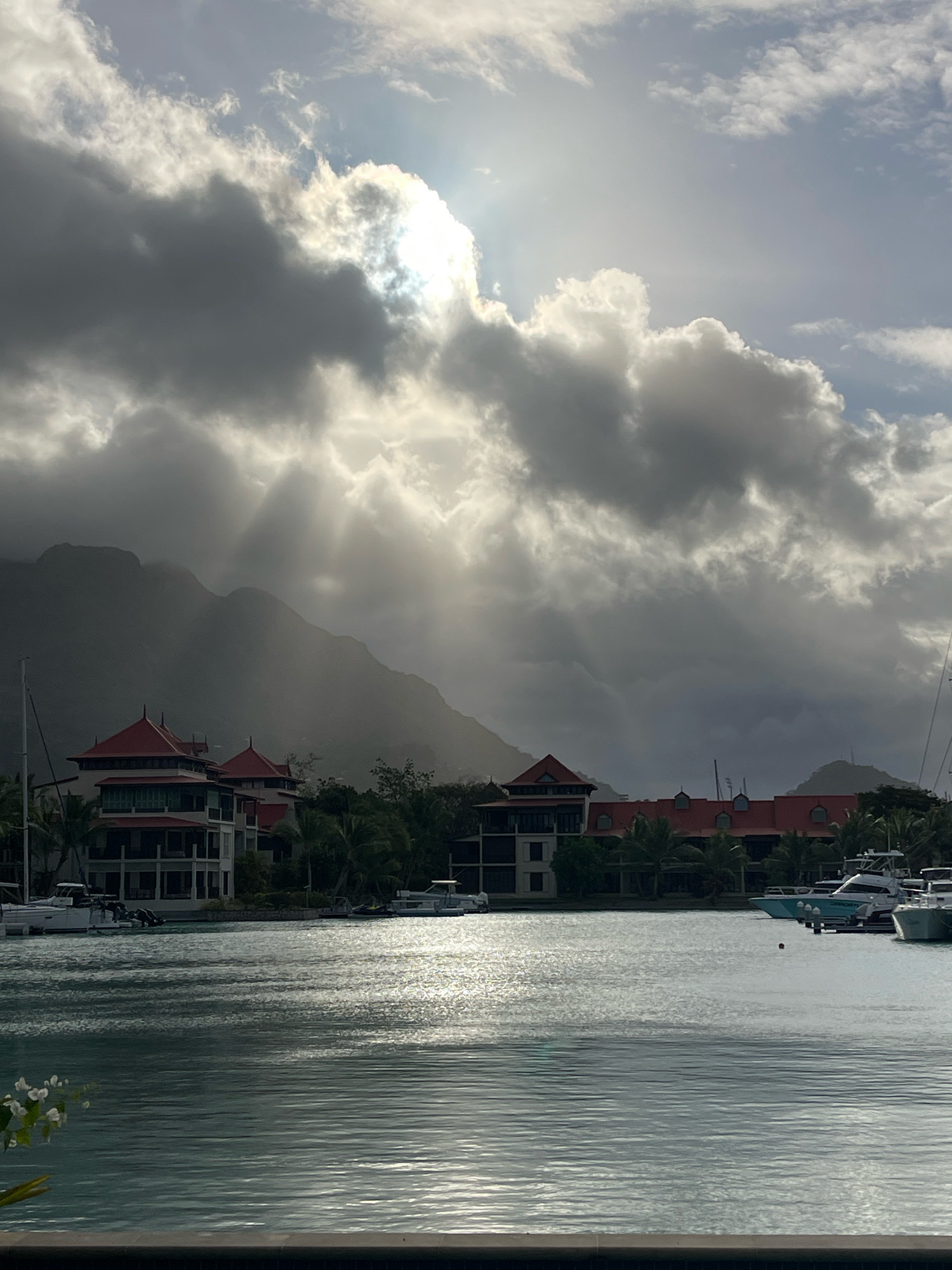 The image shows a body of water in front of a waterfront property in Providence, Seychelles. The water is calm and reflects the light from the sun, which is partially obscured by dramatic, dark clouds. Sunbeams are breaking through the clouds and illuminating the scene. On the shore, there are several multi-story buildings with red tiled roofs, interspersed with palm trees. Several boats are docked in the water, including a white sailboat on the left and a white motor yacht on the right. In the background, a mountain range is visible, shrouded in mist and clouds. The overall mood is serene and somewhat dramatic due to the lighting conditions.