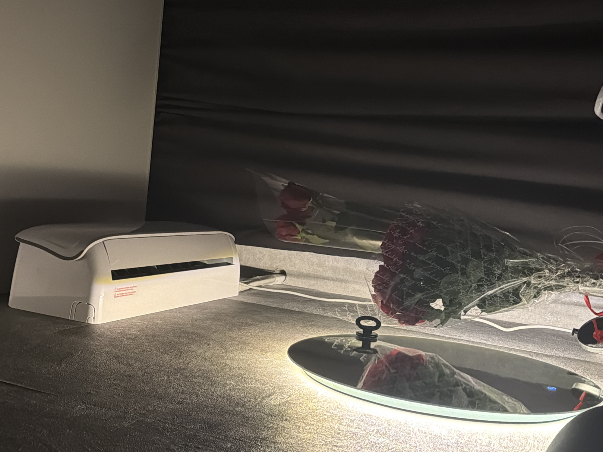 A bouquet of red roses wrapped in clear plastic sits on a table. The roses are in front of a mirror with a lit edge and a key hanging on it. Above the mirror, an air conditioner unit is mounted on a wall. To the right of the roses, dark curtains are visible. The scene is indoors, in an environment that could be a bedroom or living room. The overall lighting is dim, with the mirror and air conditioner providing artificial light sources. The photograph is taken from a low angle. There are no people present in the image.