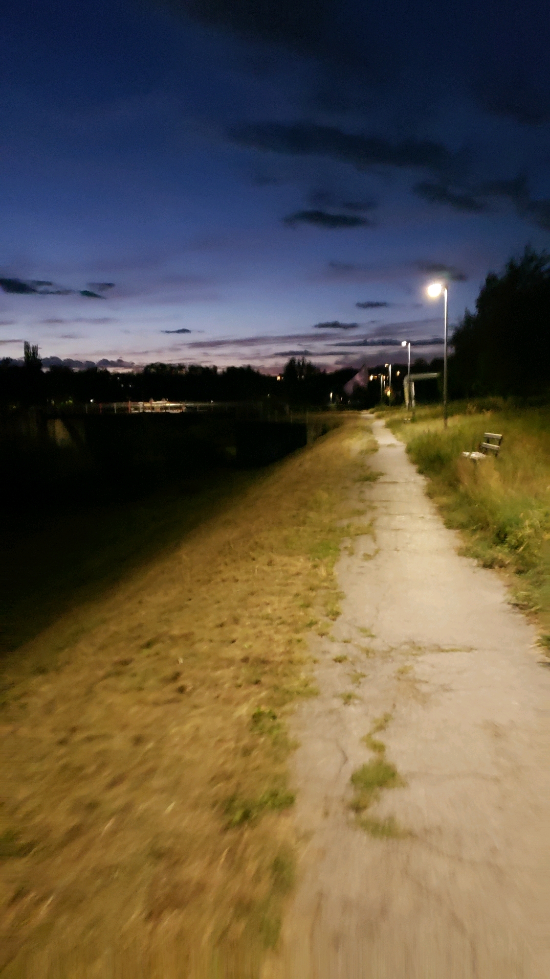 A pathway runs alongside a dark, grassy embankment, leading towards a streetlamp illuminating the scene. The sky above is a deep blue, with streaks of orange and purple from the setting sun, and dark clouds are scattered across it. In the distance, trees and buildings are silhouetted against the twilight sky. A wooden bench is visible on the right side of the pathway. The overall impression is one of a peaceful, natural setting within a city. The image was captured in Prešov, Slovakia.