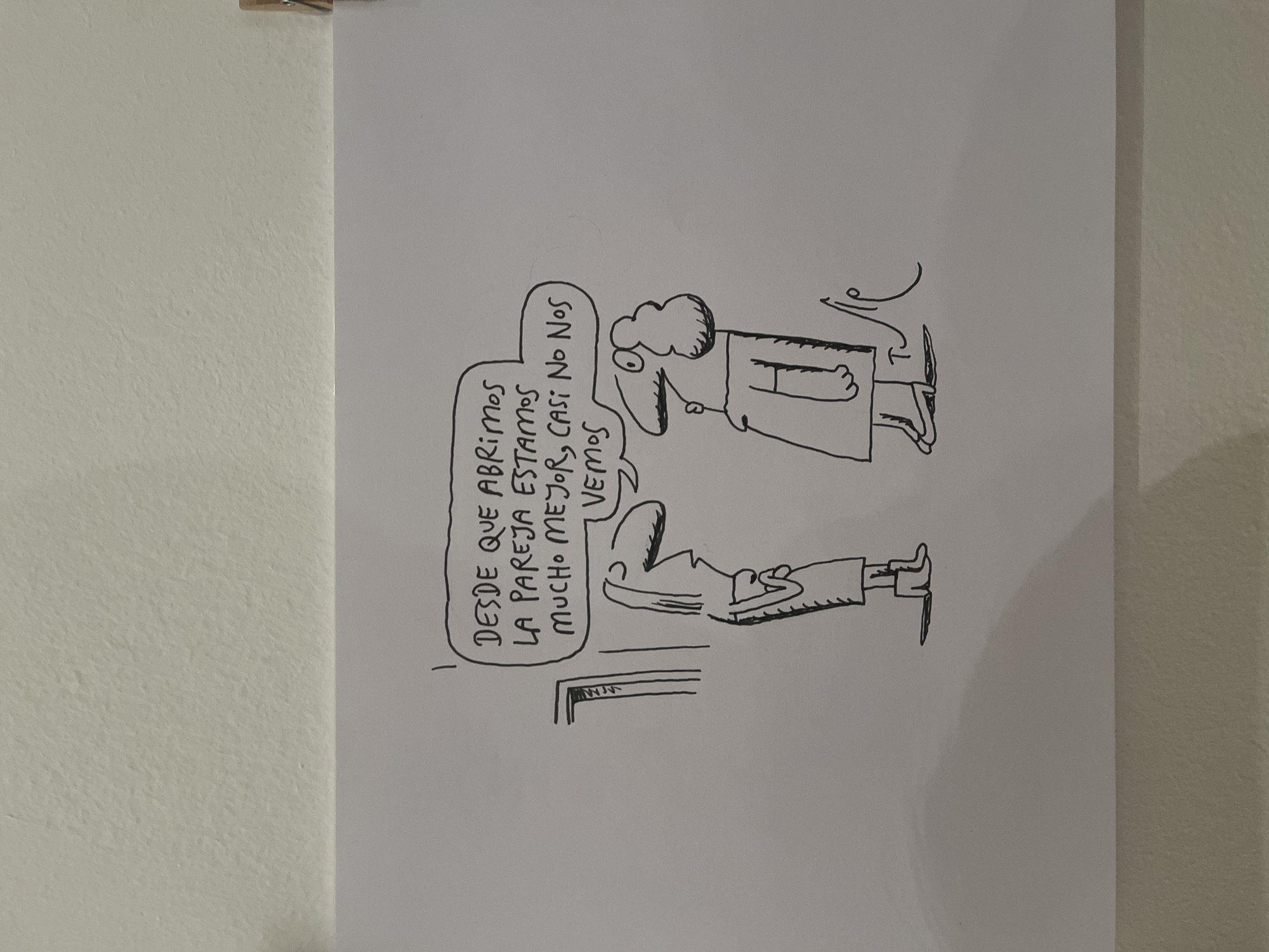 A hand-drawn cartoon on a white paper shows two men, presumably a couple, standing on either side of a doorway. One man has a large nose and a puff of hair, the other has a long nose and is shorter. A speech bubble above the shorter man says, "Desde que abrimos la pareja estamos mucho mejor, casi no nos vemos." (Since we opened up the relationship, we are much better, we almost don't see each other). The cartoon is likely located in Buenos Aires, Argentina, given the Spanish language used in the speech bubble. The image suggests a commentary on open relationships.