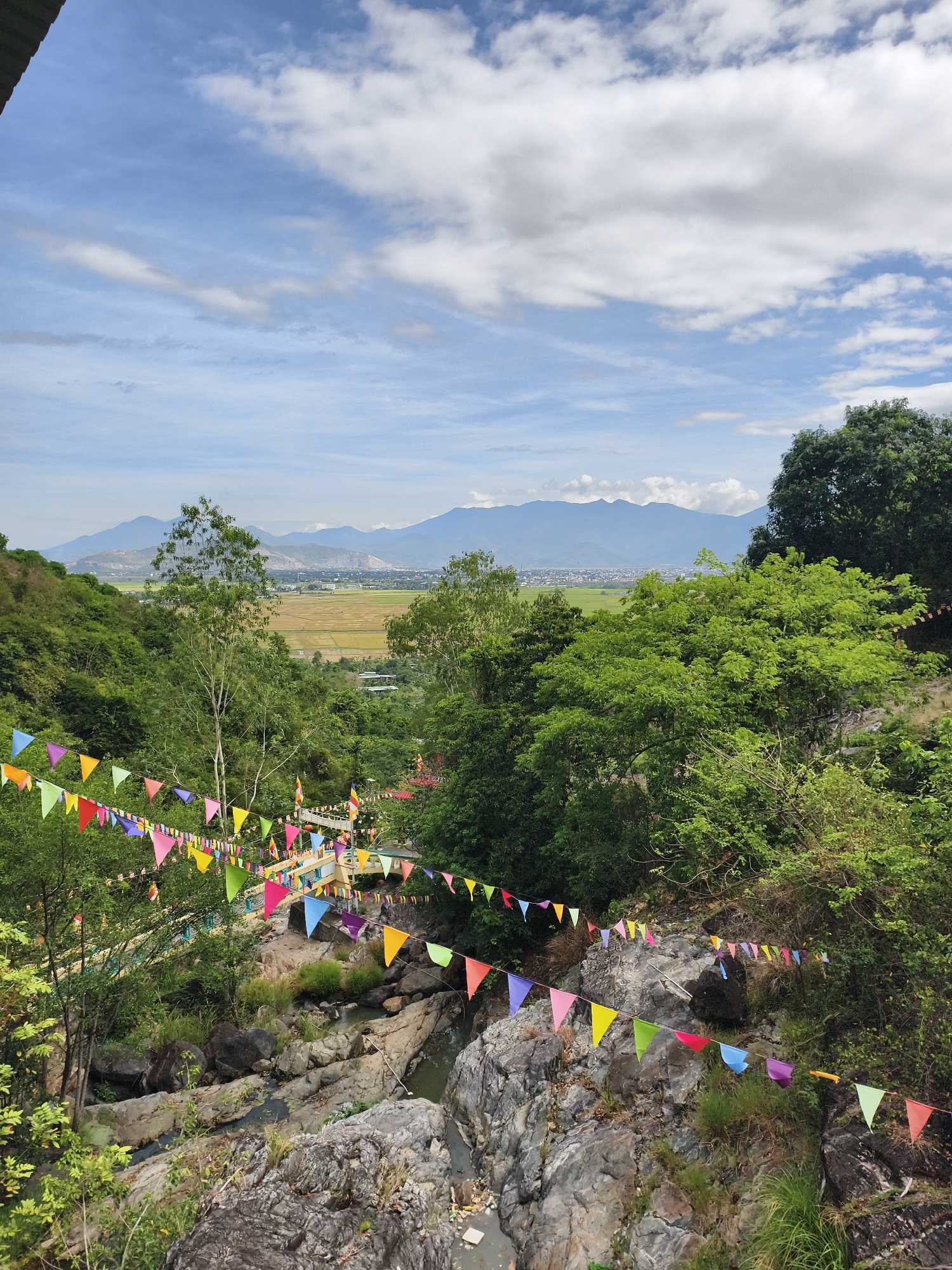The photograph captures a landscape scene in Tay Nha Trang Ward, Vietnam. The foreground features a rocky creek bed with water flowing through it. The creek bed is decorated with strings of colorful triangular flags.
Beyond the creek bed, lush green vegetation covers the hillsides. A bridge with yellow railings is visible amidst the trees. In the distance, a valley opens up with agricultural fields and some buildings. Mountain ranges form a backdrop against a sky filled with puffy white clouds and patches of blue sky.