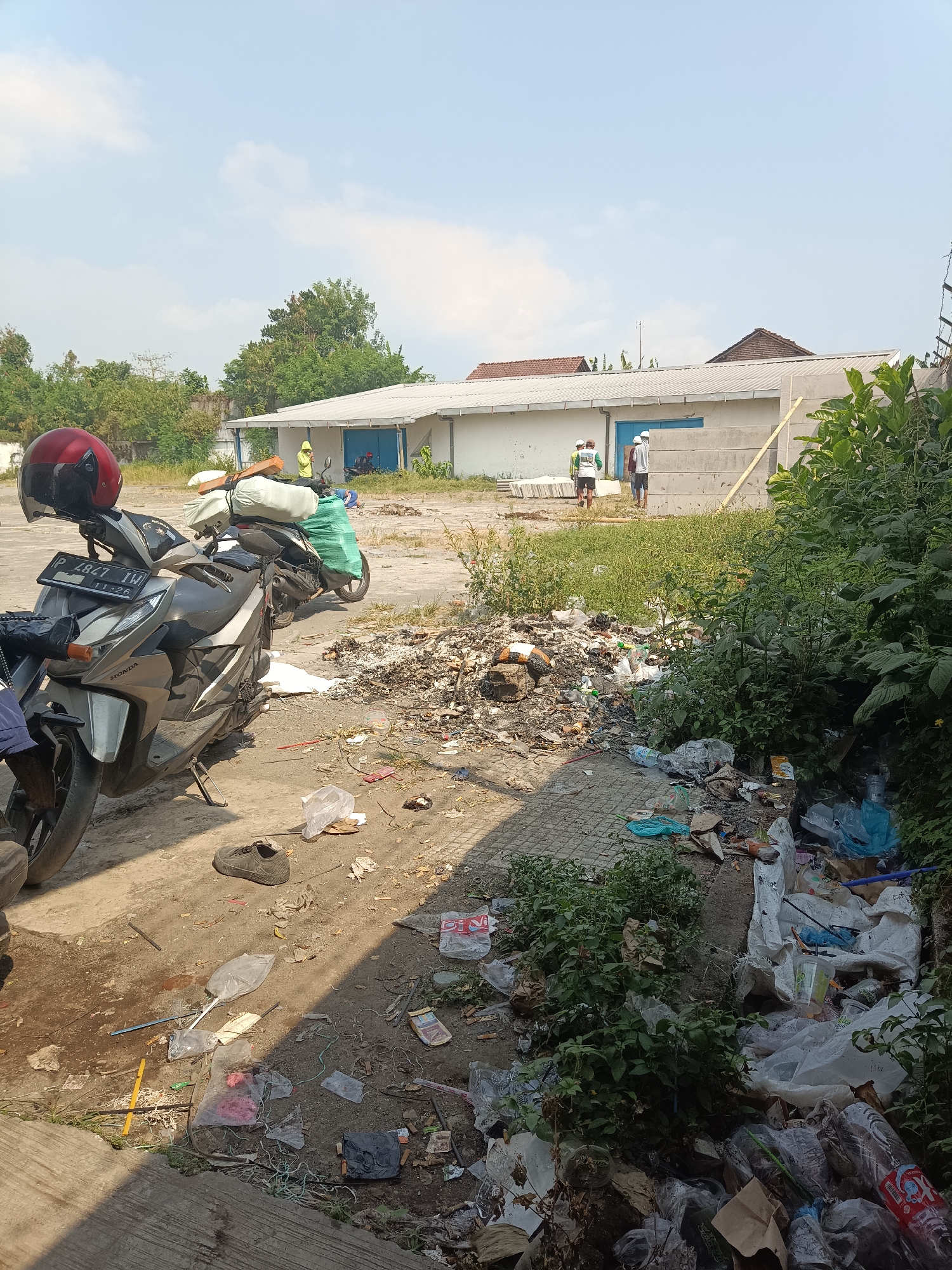 This image depicts an outdoor scene in Jember, Indonesia. In the foreground, a parked motorcycle with a red helmet on its handlebars is visible. The ground around the motorcycle and extending into the midground is heavily littered with trash, including plastic bags, bottles, and other debris. In the background, there is a white building with blue doors, and several individuals are standing near it. Some are wearing what appear to be work clothes, and one person is wearing a yellow hooded jacket. The environment is sunny with a clear blue sky and some clouds, suggesting daytime. Lush green foliage is present on the right side of the image, contrasting with the discarded waste. The overall impression is of a somewhat unkempt outdoor area, possibly a service entrance or staging ground for the building.