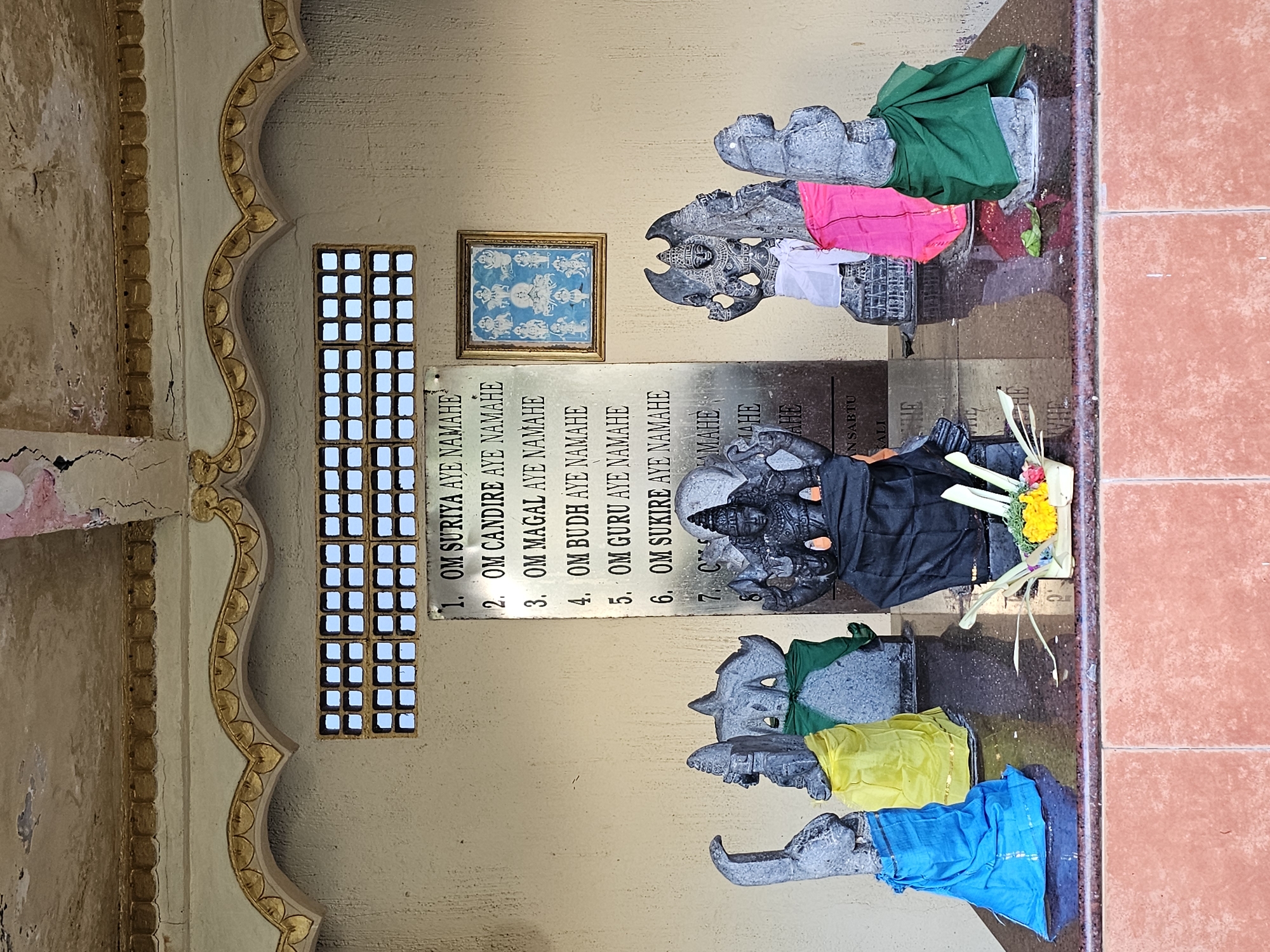 A frontal, medium shot depicts a religious shrine in what appears to be Denpasar, Indonesia. The shrine is composed of a beige painted wall with a lattice window, a framed picture, and a plaque with names. Six stone carvings are partially covered in cloth and sit in the foreground. A series of texts are placed next to the statues, and a piece of religious art lies on the right corner of the middle carving. The background is an earthy, light-yellow color, while the statues in the foreground stand on a reddish tile.