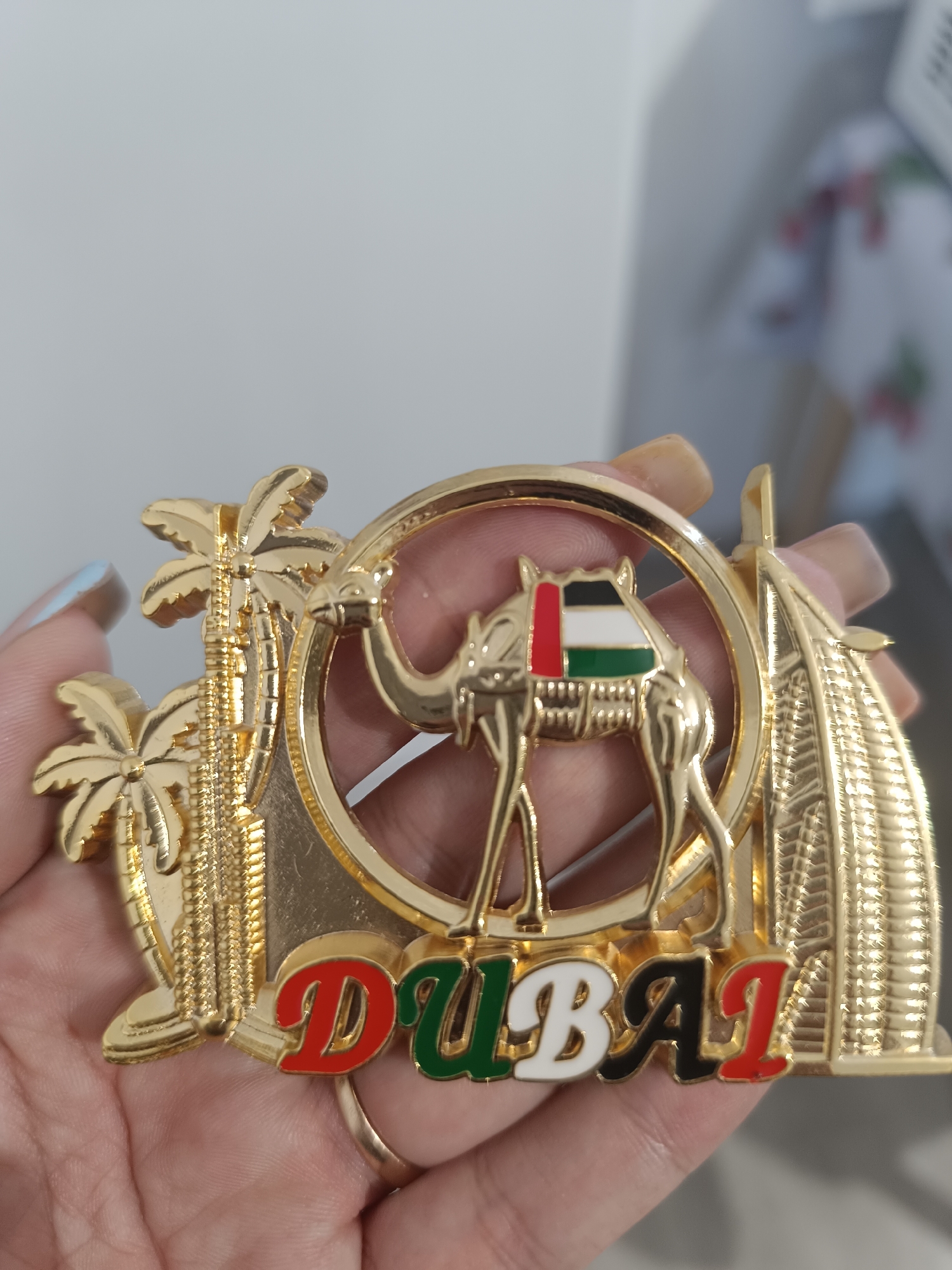 A gold-colored souvenir magnet is being held by a person's hand. The magnet is shaped like a scene with several iconic elements. In the center, a camel is depicted wearing a saddle with the colors of the UAE flag. Behind the camel is a circular frame. To the left of the camel, there are palm trees and what appears to be a modern skyscraper. To the right, a building resembling the Burj Al Arab is visible. Below these elements, the word "DUBAI" is written in colorful lettering: red for "D", green for "U", white for "B", black for "A", and red for "I". The background is a plain, light gray wall. The image does not show or depict any location in the city of Taraz city administration, Kazakhstan.