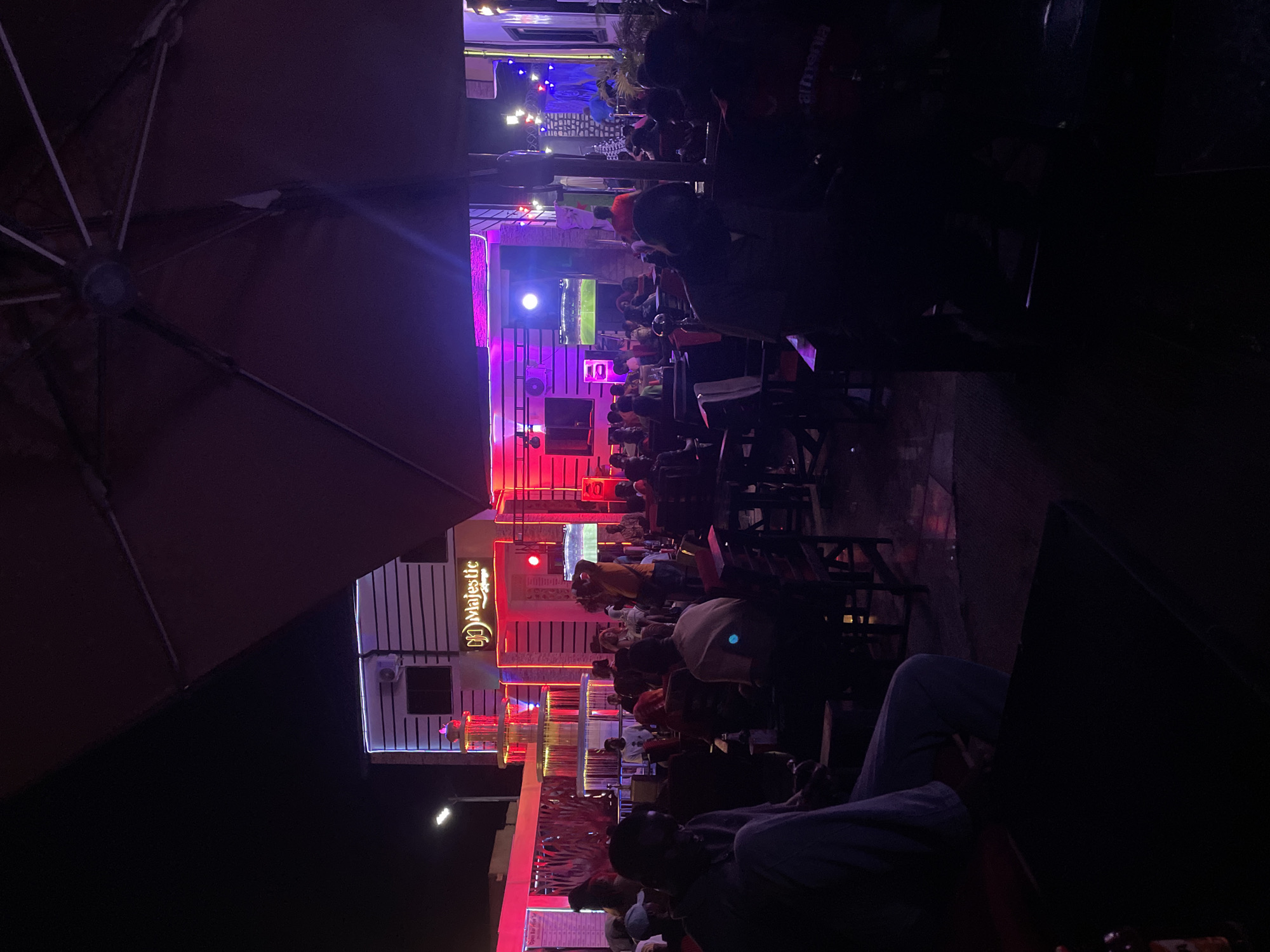 The image depicts an outdoor gathering in Tumfure, Nigeria, possibly at a restaurant or bar called "Majestic," as indicated by the sign on a white multi-story building. The building is adorned with vibrant red, purple, and blue neon lighting. People are seated at tables and chairs, many facing a screen displaying a soccer game. The area is dimly lit, with an umbrella partially visible in the foreground. The lighting and activity suggest it is nighttime. Some individuals are clearly visible while others are obscured by shadows. A person in the lower right corner is wearing light-colored pants and a grey shirt.