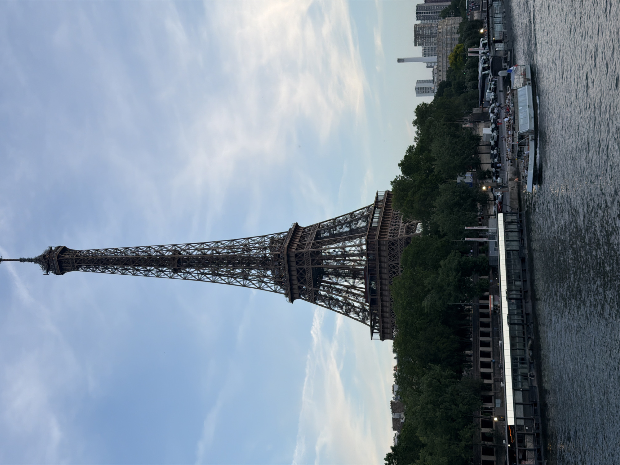 The image shows the Eiffel Tower in Paris, France, as viewed from a boat or bridge on the Seine River. The tower is positioned on the left side of the frame, reaching towards the sky. To the right of the tower, a line of trees and buildings runs along the riverbank, leading to a pier where tour boats are docked. The Seine River flows in the foreground, with its surface reflecting the surrounding lights and structures. The sky is pale blue with scattered white clouds.