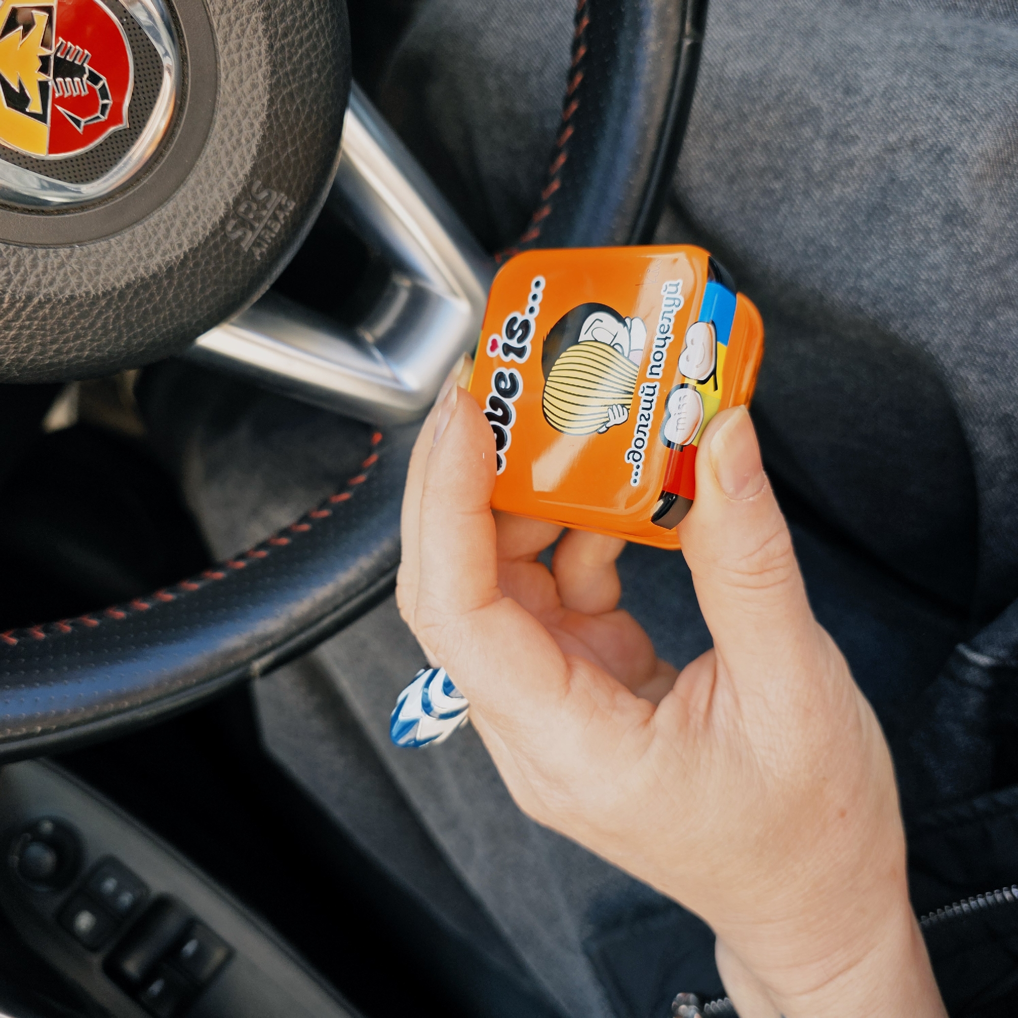 A person is holding an orange candy box in their right hand inside a car. The candy box has a cartoon illustration of a boy and girl with the words "love is..." written on it in English and Russian. The steering wheel of the car is visible in the background, featuring the Abarth logo. The interior of the car, including the seat and dashboard, is also visible. The location is Saint Petersburg, Russia based on the Cyrillic text on the box.