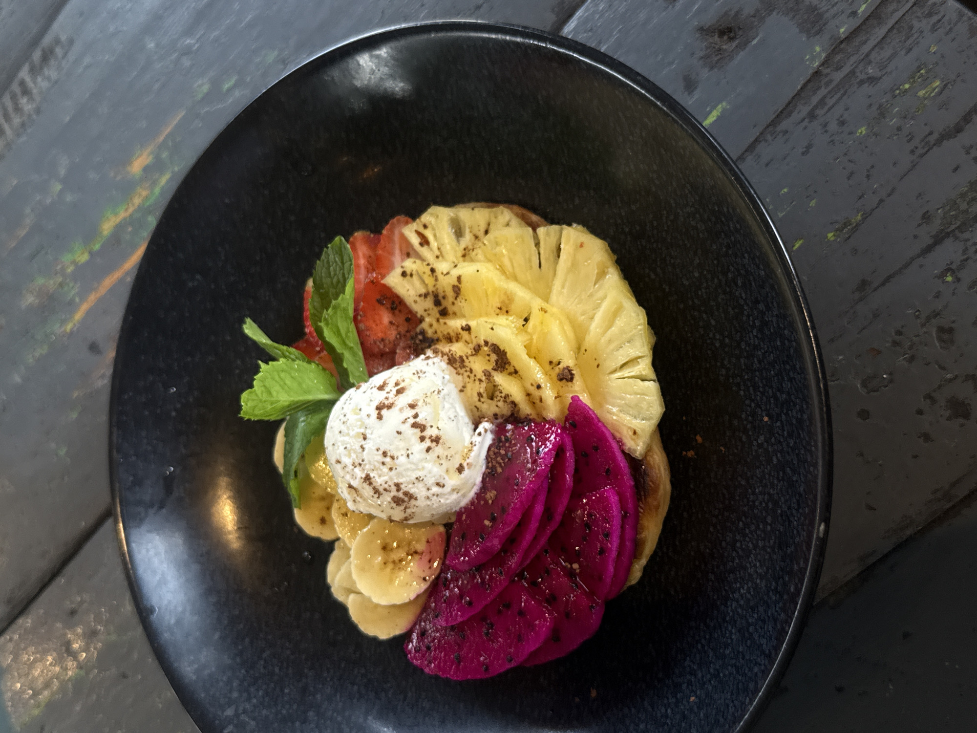 An overhead shot captures a vibrant, aesthetically arranged dessert or breakfast dish presented in a dark, textured ceramic bowl. The bowl is set on a dark, possibly wooden, table exhibiting visible grain and some subtle, greenish-yellow discolorations or reflections on its surface.
The dish itself is a colorful assembly of fresh fruits and a creamy topping. Sliced bright yellow pineapple forms an arc at the top, followed by striking magenta-purple dragon fruit slices, complete with their tiny black seeds, arranged along the right side. Pale yellow, round banana slices are placed at the bottom, and bright red strawberry slices complete the fruit circle on the left.
A generous scoop of white cream, possibly ice cream, whipped cream, or yogurt, is centered on the banana and strawberry slices. It is lightly dusted with a dark brown powder, likely cocoa or cinnamon. Several fresh green mint leaves are strategically placed among the fruits, adding a pop of color and garnish.
The overall presentation is fresh and appealing, with the vivid colors of the fruit contrasting against the dark plate and table. There is no visible text in the image. The tropical fruits and presentation style are consistent with a setting in North Kuta, Indonesia.