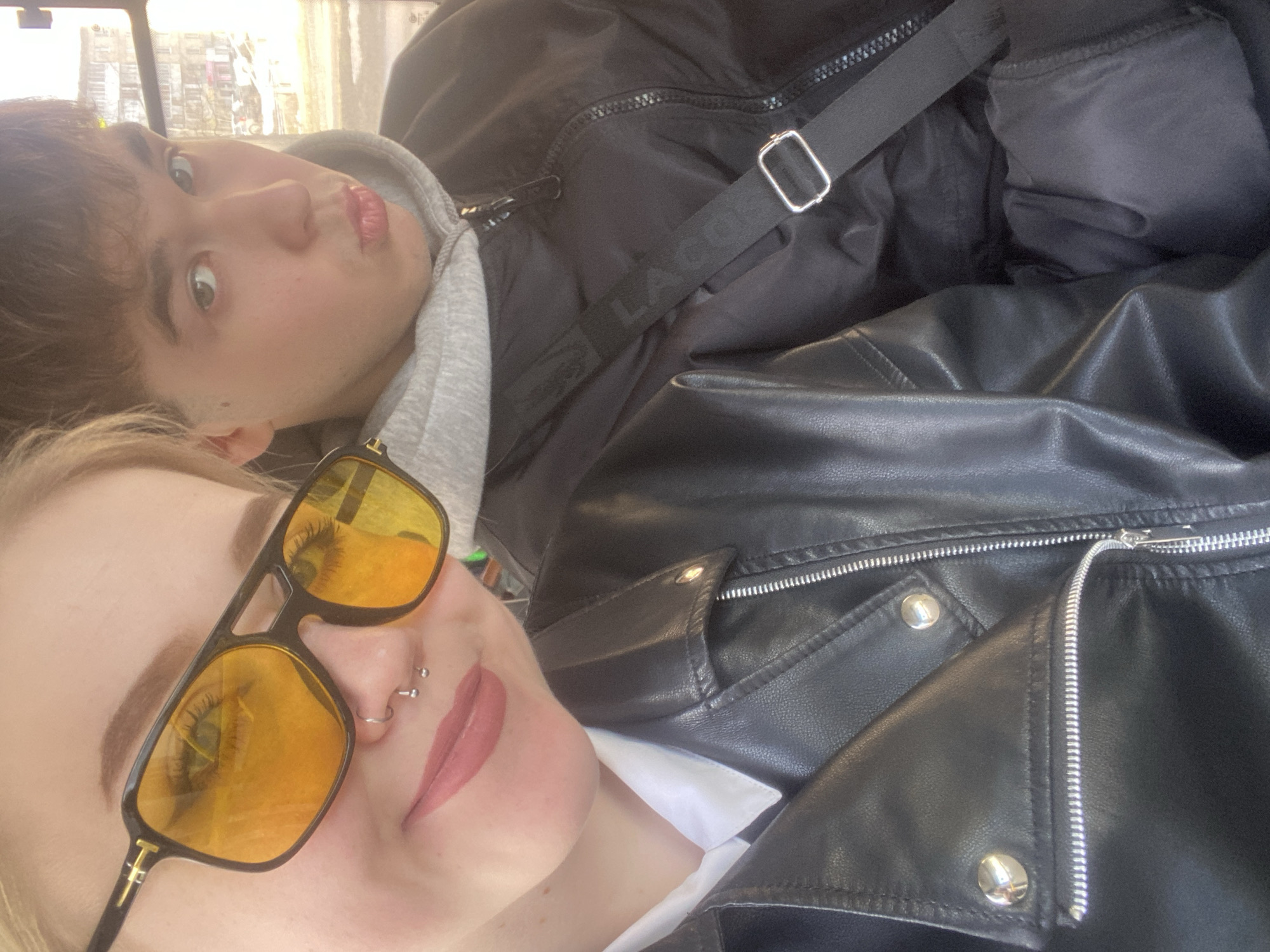 Two people are in a vehicle or indoor space. A person with blonde hair and sunglasses is in the foreground. A second person is behind them with dark hair. The first person is wearing a black leather jacket and has a double nose piercing. The second person is wearing a dark jacket and has a Lacoste bag strap across their chest. The location is not discernible.