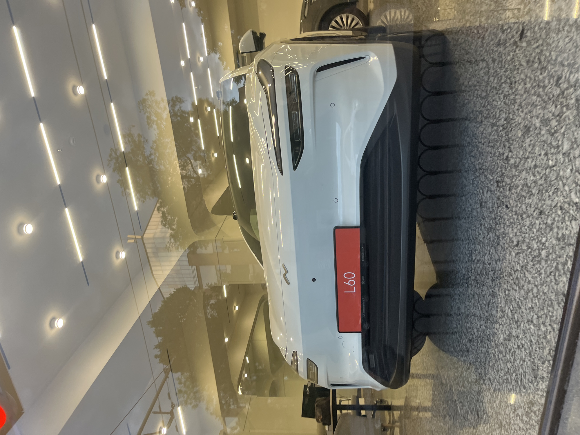The image shows a white NIO L60 electric vehicle, possibly a showroom model, parked indoors in what appears to be a dealership or exhibition space. The car has a red license plate with "L60" printed in white. The location is likely Yerevan, Armenia. The surrounding environment includes a polished floor and large windows or glass walls, allowing outside light and reflections of the interior. Artificial lighting fixtures are visible on the ceiling. A portion of another vehicle is visible in the background. The photograph is taken from a medium angle, looking at the rear of the L60, showcasing the rear lights, bumper, and license plate.
