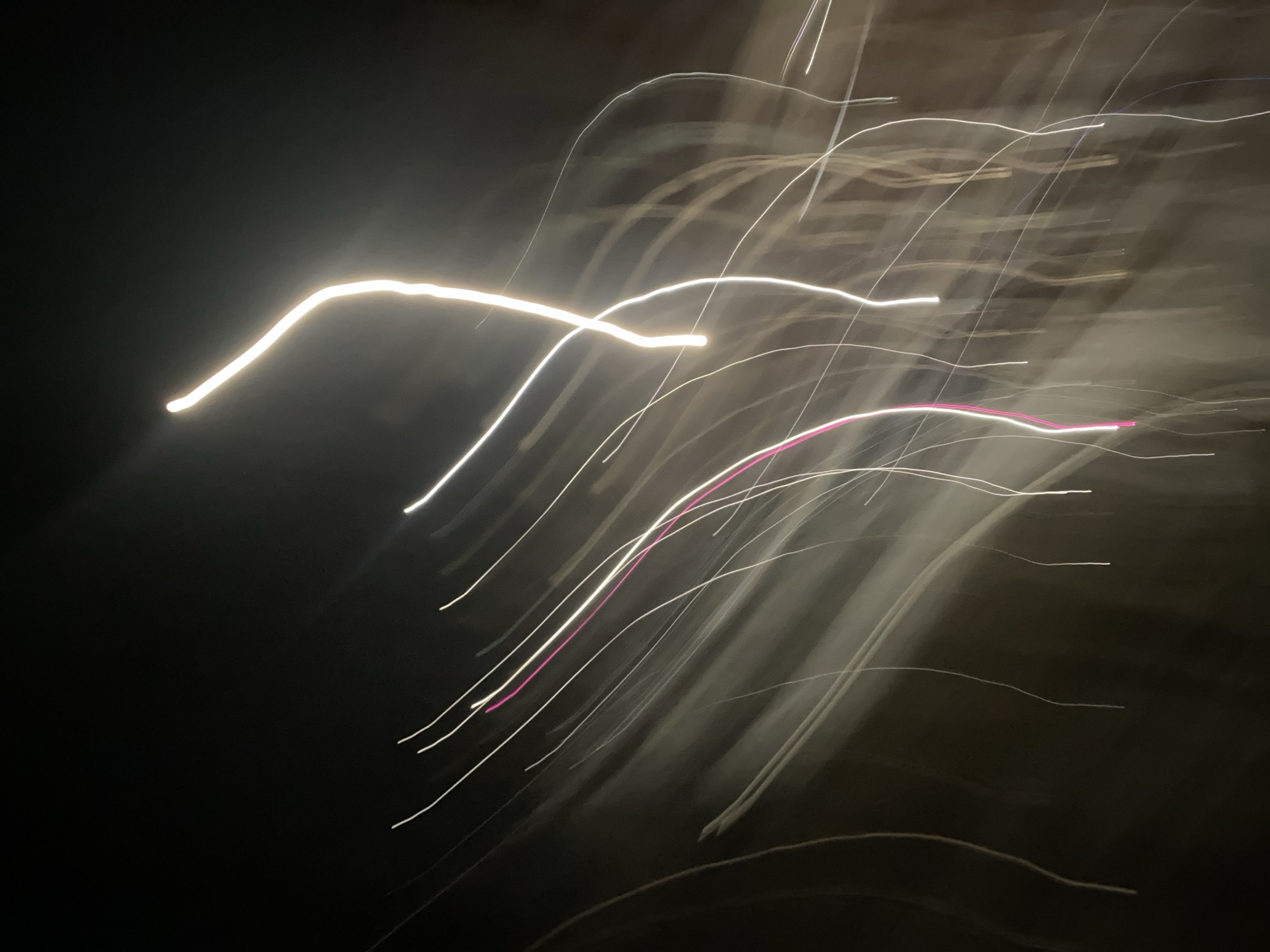 The image is a long exposure photograph taken at night. Several streaks of light, primarily white and a few pink, are visible against a dark background. The light streaks are curved and appear to be moving from the upper right to the lower left of the frame. The setting is likely an urban environment, given the presence of artificial lights. The location of the image in the city of Chaikovsky, Russia is specified by the user. Further details are unknown.