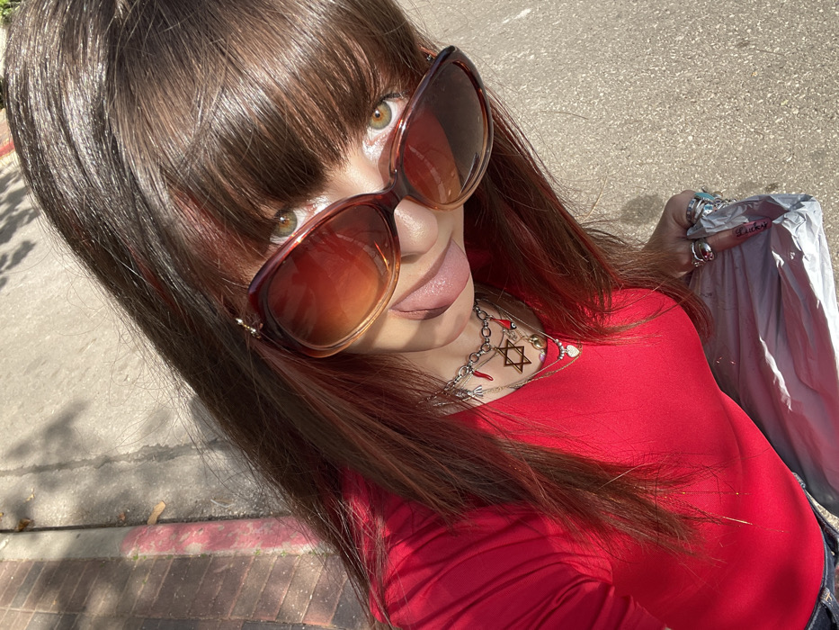 A high-angle, close-up photograph depicts an individual outdoors in the city of Raanana, Israel, under bright sunlight. The subject, appearing female, has long, dark brown hair with reddish highlights and bangs. They wear large, brown-framed, gradient-lensed sunglasses, revealing greenish-brown eyes above the frames. The tongue is extended from the mouth. The individual is dressed in a red long-sleeved top. Multiple layered silver-toned necklaces are visible, featuring charms including a prominent Star of David, a red chili pepper, and an evil eye. Thinner chains with small pendants, including a heart, are also present. The right hand, adorned with several silver-toned rings, holds an object wrapped in silver-colored foil. A pink inscription is visible on a label affixed to the foil. The background consists of a light-colored concrete or asphalt sidewalk with shadows, and a red-painted curbstone is visible to the upper left.