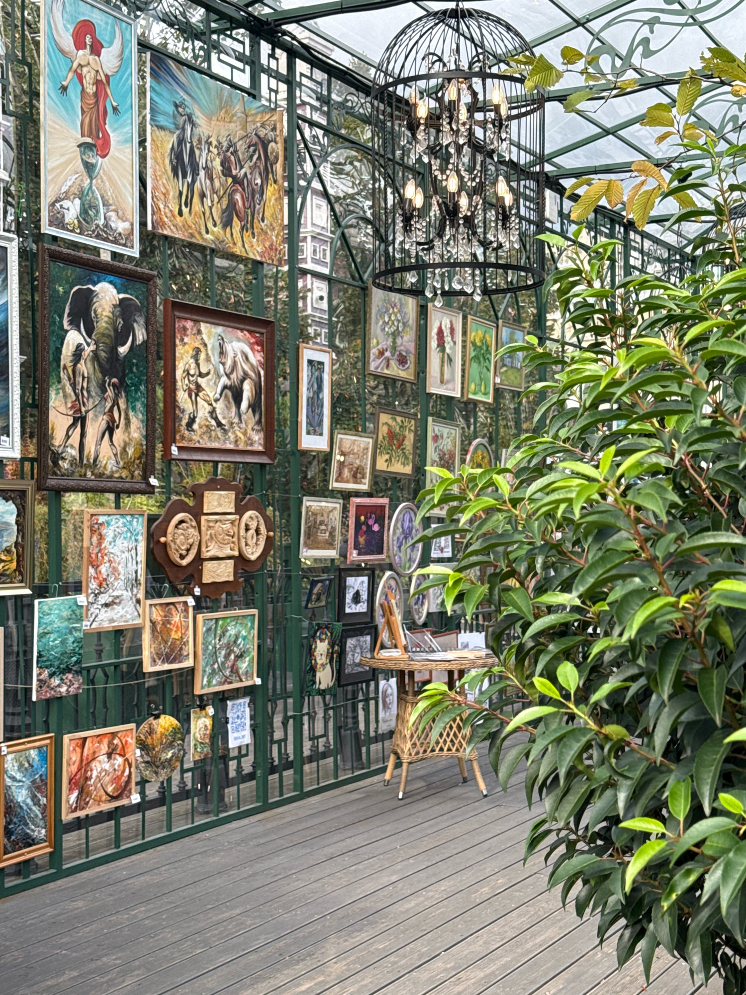 The image depicts an outdoor art exhibition, likely in Moscow, Russia, characterized by a collection of paintings and artworks displayed along a green metal fence within a greenhouse-like structure with a glass roof. Prominently featured are several framed paintings, including a large artwork on the left with a winged figure over an hourglass, a painting of horses and riders, and another depicting an elephant and figures. Smaller framed pictures and circular artworks are also visible, showcasing a variety of subjects from floral arrangements to abstract designs. In the background, a large black birdcage-like chandelier with multiple lights and crystal accents hangs from the ceiling. A small wicker table with some papers and a wicker chair are also present, suggesting a relaxed viewing area. Lush green foliage from a large plant partially obscures the right side of the image. The ground is made of wooden planks. No people are visible in the scene.