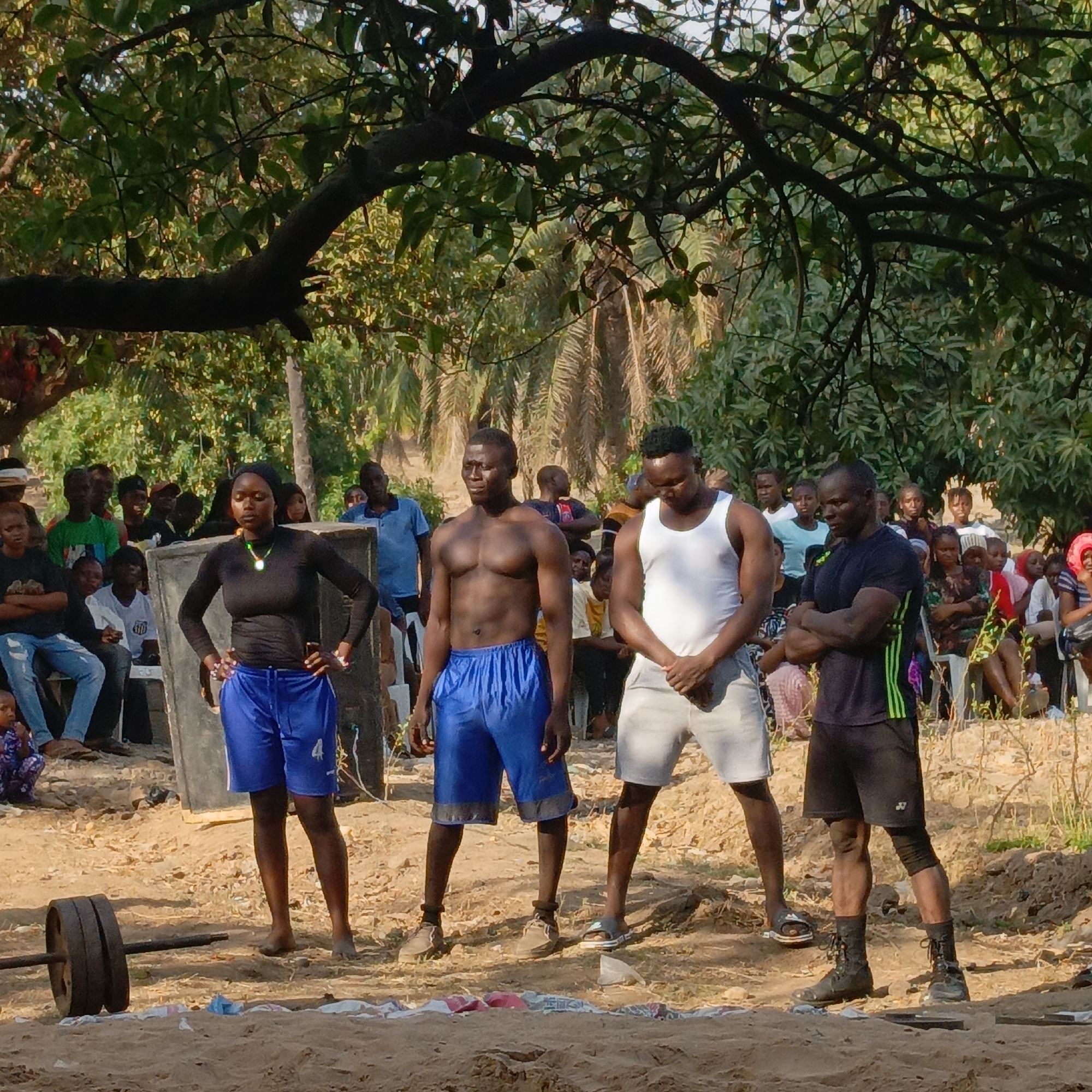 An outdoor scene in Akwanga, Nigeria, depicts four individuals positioned in the foreground of a gathering. From left to right: A female subject stands with hands on her hips, wearing a black long-sleeved top, blue athletic shorts, and a dark head covering, with a multi-colored beaded necklace. She is barefoot. Next, a male subject with a muscular physique stands bare-chested, wearing blue athletic shorts, also barefoot. To his right, another male subject wears a white tank top and light grey shorts, with hands clasped in front and head slightly bowed; he wears dark slides. The fourth male subject stands with arms crossed, wearing a black short-sleeved shirt with green vertical stripes on the side, black shorts, and black combat-style boots.
In the immediate foreground to the left, a barbell with two dark circular weights is resting on the sandy ground. Scattered pieces of light-colored fabric or plastic are visible on the ground between the subjects. The ground surface is predominantly loose, light brown dirt or sand.
Behind these four individuals, a diverse crowd of approximately 50-70 spectators is visible, consisting of adults and children. Many spectators are seated on white plastic chairs or standing, observing the scene. The background features dense green foliage, including palm trees and other broadleaf trees, under a bright sky. A large, dark tree branch extends horizontally across the upper portion of the frame, partially obscuring the sky and upper canopy. The overall lighting suggests daylight conditions.