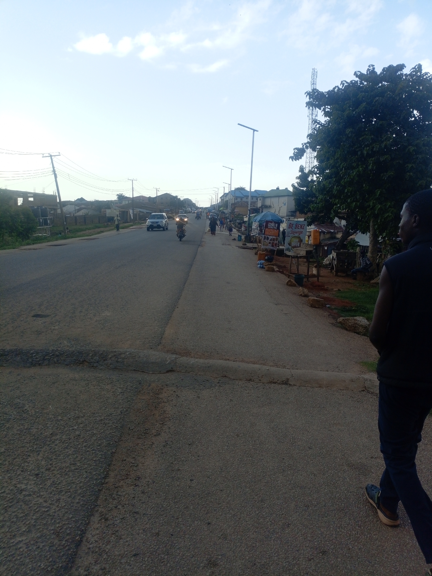The image depicts a street scene in Jos, Nigeria, possibly during the late afternoon or early evening. The road is paved and appears to be a moderately busy thoroughfare with vehicles, including cars and a motorcycle, moving along it. On the right side of the road, there are several informal businesses or stalls set up, indicated by structures with awnings and signage, one of which reads "DE BENZ Lounge." A person is walking on the sidewalk on the right side of the street, facing away from the camera. In the background, there are buildings and trees, and a telecommunications tower is visible in the distance. The sky is partly cloudy, suggesting a typical weather condition for the region. The overall impression is of a lively urban environment with commercial activity lining the street.
