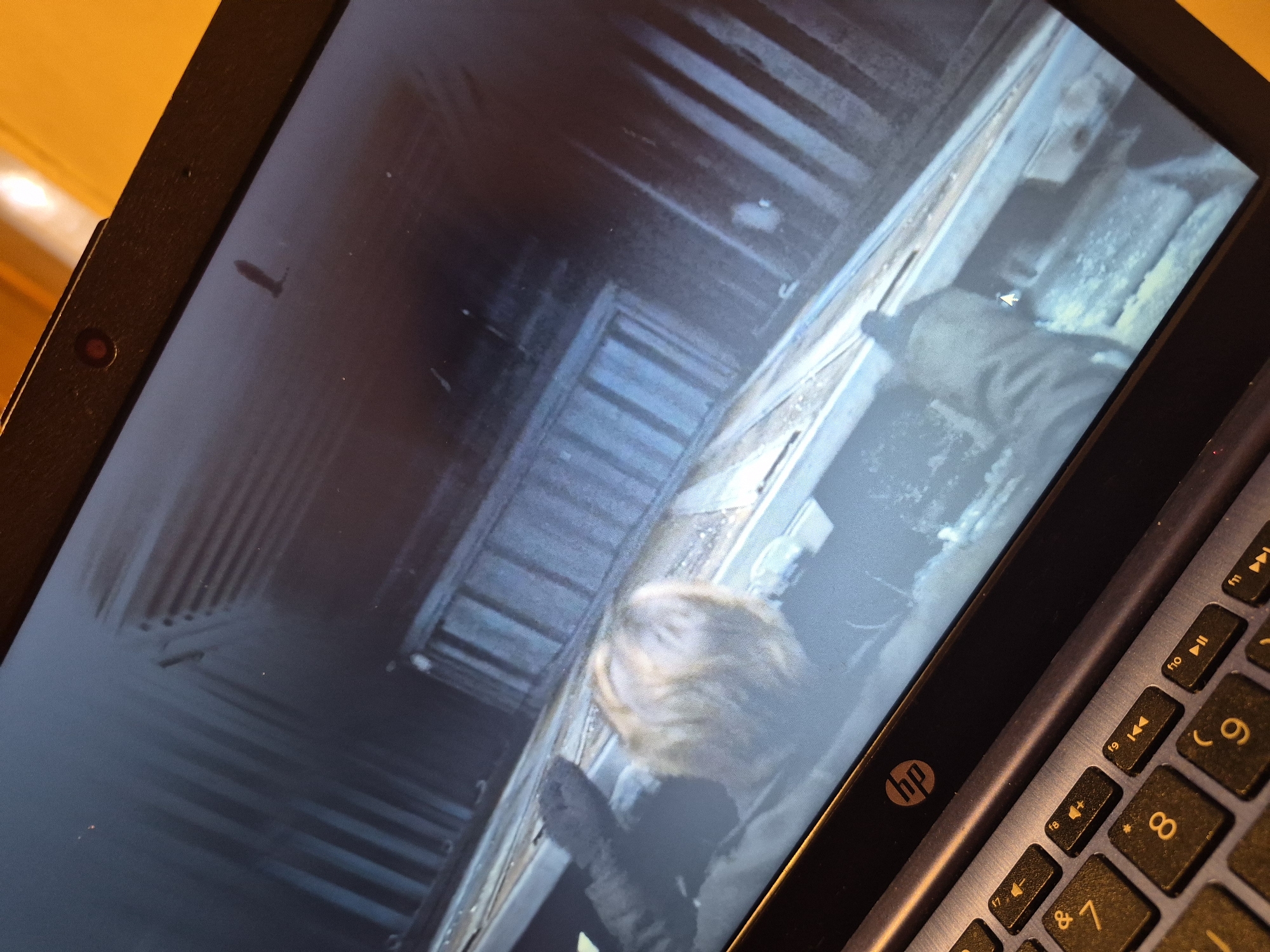 The image shows a laptop screen displaying a video game scene. The game depicts a first-person perspective looking down into a narrow, industrial-looking space, possibly a ventilation shaft or a similar confined area. A character's blond hair and gloved hands are visible at the bottom of the screen, suggesting they are either climbing or descending. The environment appears dark and metallic, with visible structural elements like beams and grates. The laptop itself is an HP model, indicated by the logo on the bezel above the keyboard. The keyboard is partially visible in the foreground. The scene does not provide any specific details about the city of Sandwell, United Kingdom.