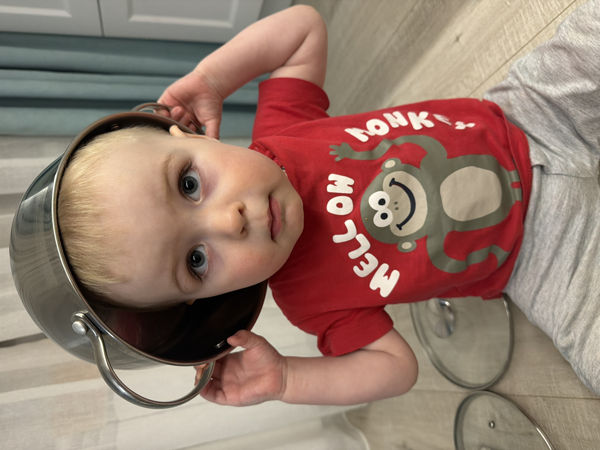 A fair-skinned child with blond hair is holding a metal pot over his head while lying on a light wood floor. He is wearing a red t-shirt with a monkey design and grey sweatpants. The background includes a blue curtain or wall. There is no evidence of the location being Votkinsk, Russia. There are also metal pot lids on the floor.