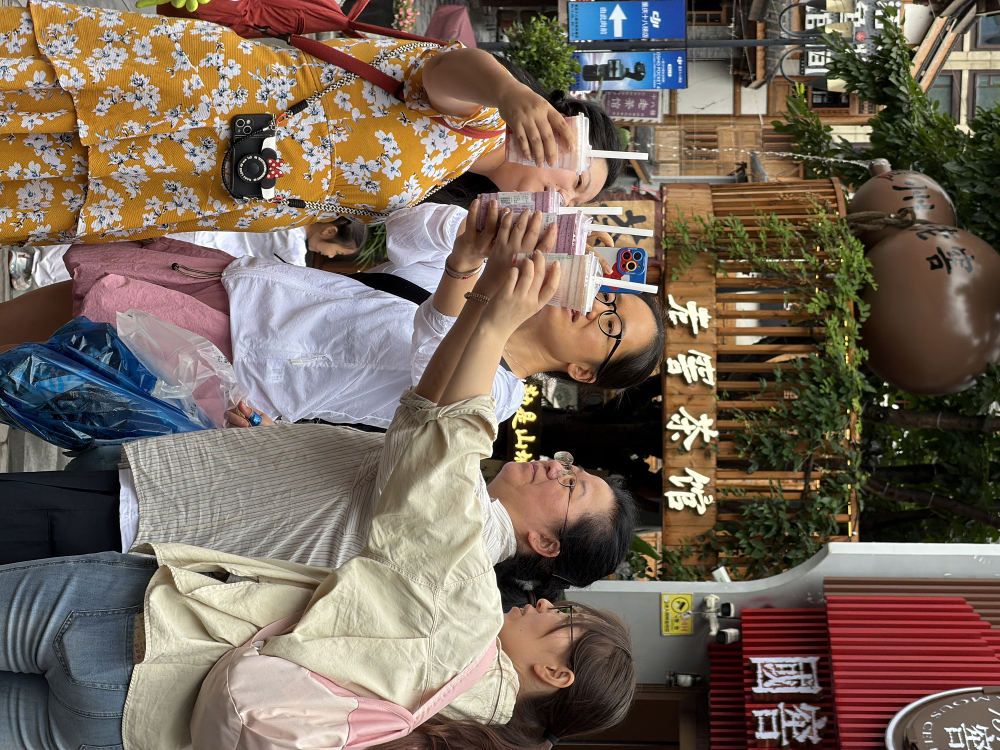 In Yuzhong District, China, four women are standing on a street, holding drinks. The woman on the far left wears a yellow floral dress and a black camera-shaped purse. The second woman from the left, wearing a white shirt and pink skirt, is holding a phone. The third woman from the left, in a striped shirt, also holds a drink. The woman on the far right, in a tan jacket and jeans, has a pink backpack. Behind them, a wooden sign with Chinese characters for "Lao Chun Tea House" is visible, along with large decorative round objects, greenery, and building facades. The scene appears to be in a commercial area with pedestrian traffic.