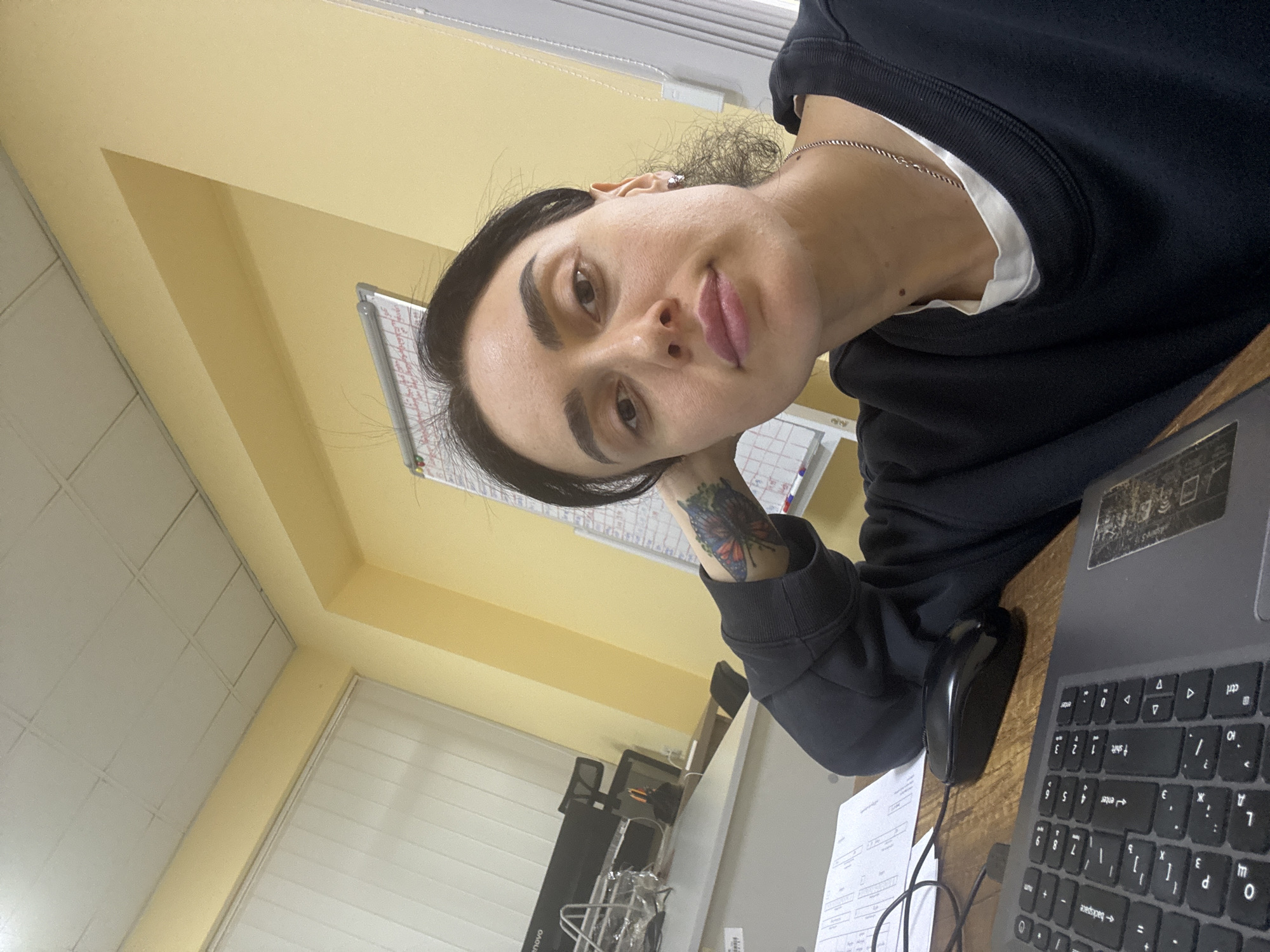 A person with dark hair and eyebrows, wearing a dark sweatshirt and a silver chain, is sitting at a desk with a laptop, mouse, and papers. The person has a tattoo on their left forearm. The background includes a whiteboard, yellow walls, and a window. A small piece of equipment is located in the lower left of the frame. The image appears to have been taken indoors. There is not enough information to determine if this is Tbilisi, Georgia.