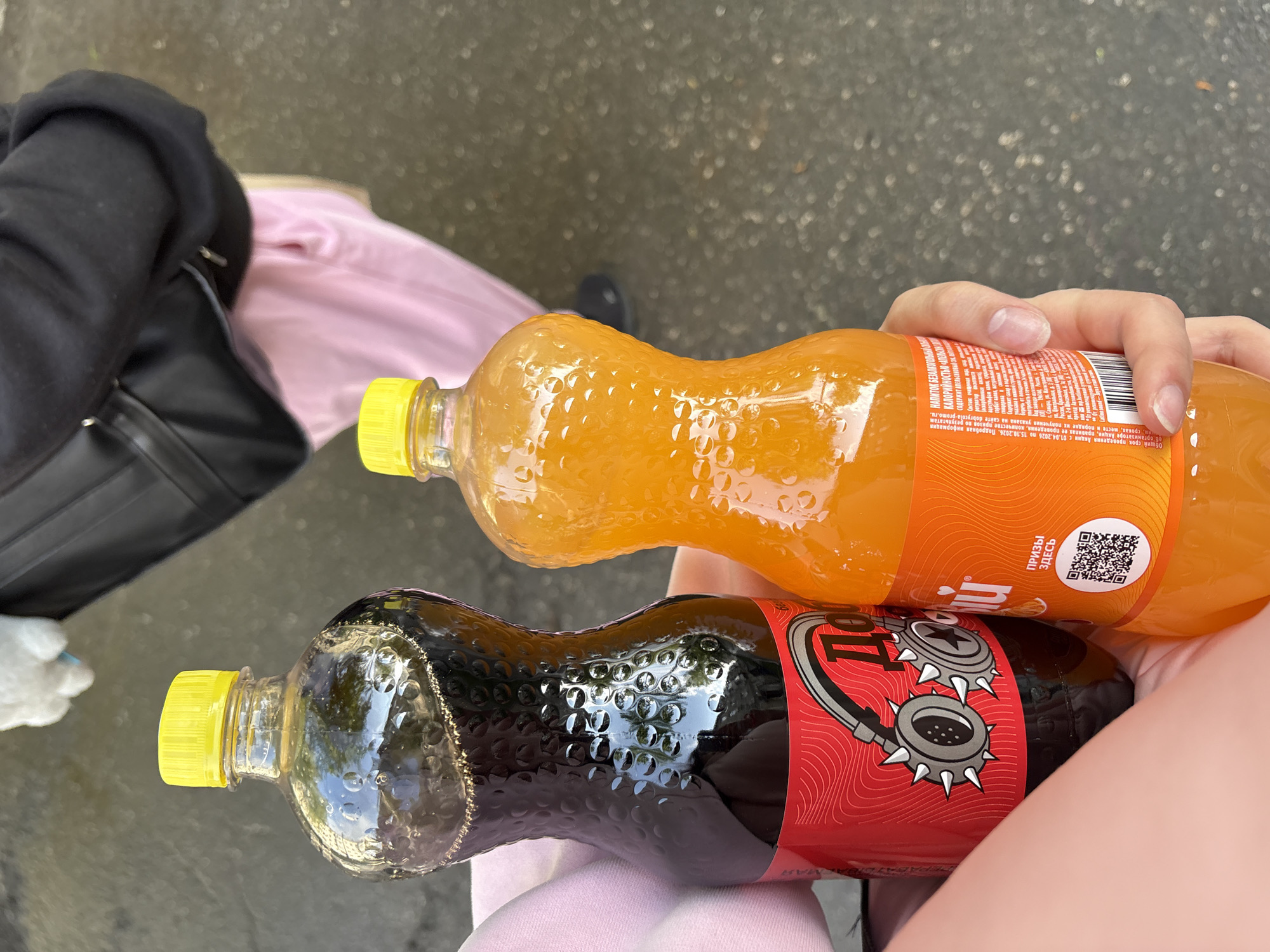 A person is holding two unopened bottles of the "Добрый" brand soft drink. One bottle contains an orange-colored liquid, likely orange soda, with a yellow cap. The other bottle contains a dark-colored liquid, possibly cola, with a yellow cap and a red label featuring a steampunk-inspired design with goggles. The label has text in Russian. Part of a black bag, a pink shirt, and gray pavement are also visible in the image. Based on the bottle labeling (which contains the cities of Samara and Chaikovsky), the scene is likely to be in Russia.