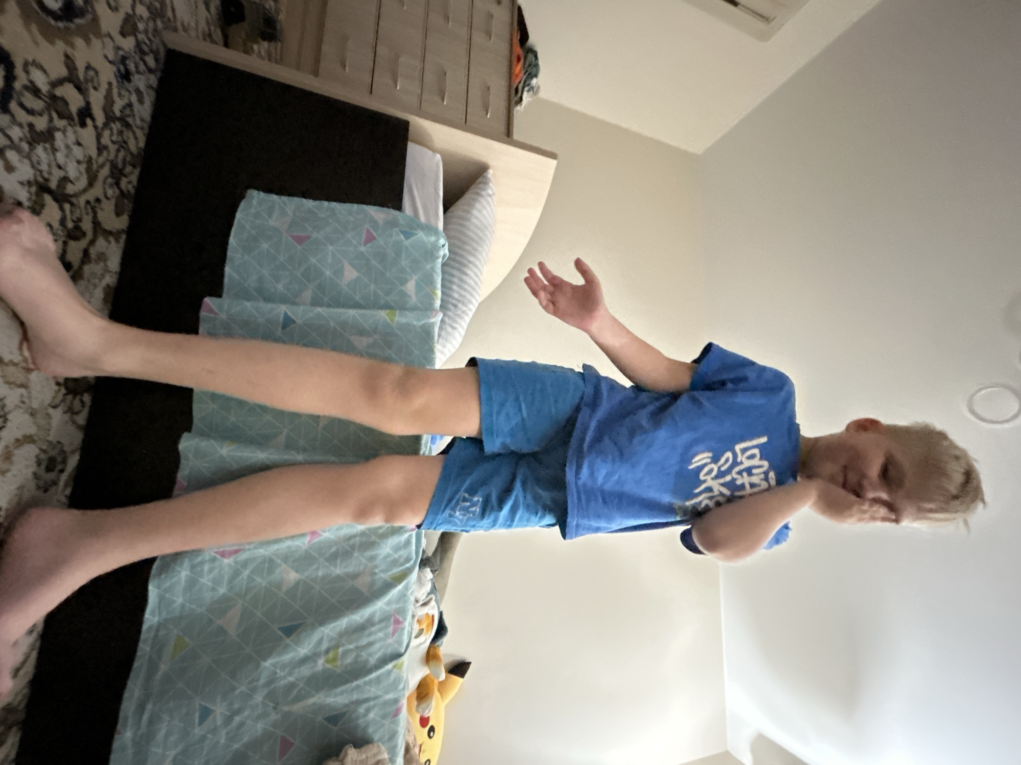 An indoor photograph, taken in Chaikovsky, Russia, captures a young boy standing in what appears to be a bedroom. The image is rotated 90 degrees clockwise. The boy, with light-colored hair, is standing barefoot on a patterned rug. He is dressed in a royal blue t-shirt featuring white, indecipherable text on the front, and matching blue shorts. His left hand is raised to partially cover his face, suggesting a shy or playful expression, while his right arm is slightly extended with an open palm.
The setting includes a bed with a light-colored wooden frame and a black headboard. The bed is covered with a light blue duvet adorned with a geometric pattern of triangles in various pastel shades. A white pillow is partially visible on the bed. Adjacent to the bed, a dark rug with intricate lighter patterns covers the floor. In the background, a light-colored wall and ceiling are visible, featuring a round recessed light fixture and an air vent. Part of a light-toned dresser or cabinet is also discernible. A yellow plush toy resembling Pikachu is tucked beside the bed near the floor. The scene depicts a casual, unposed moment, likely taken during the day with indoor lighting.