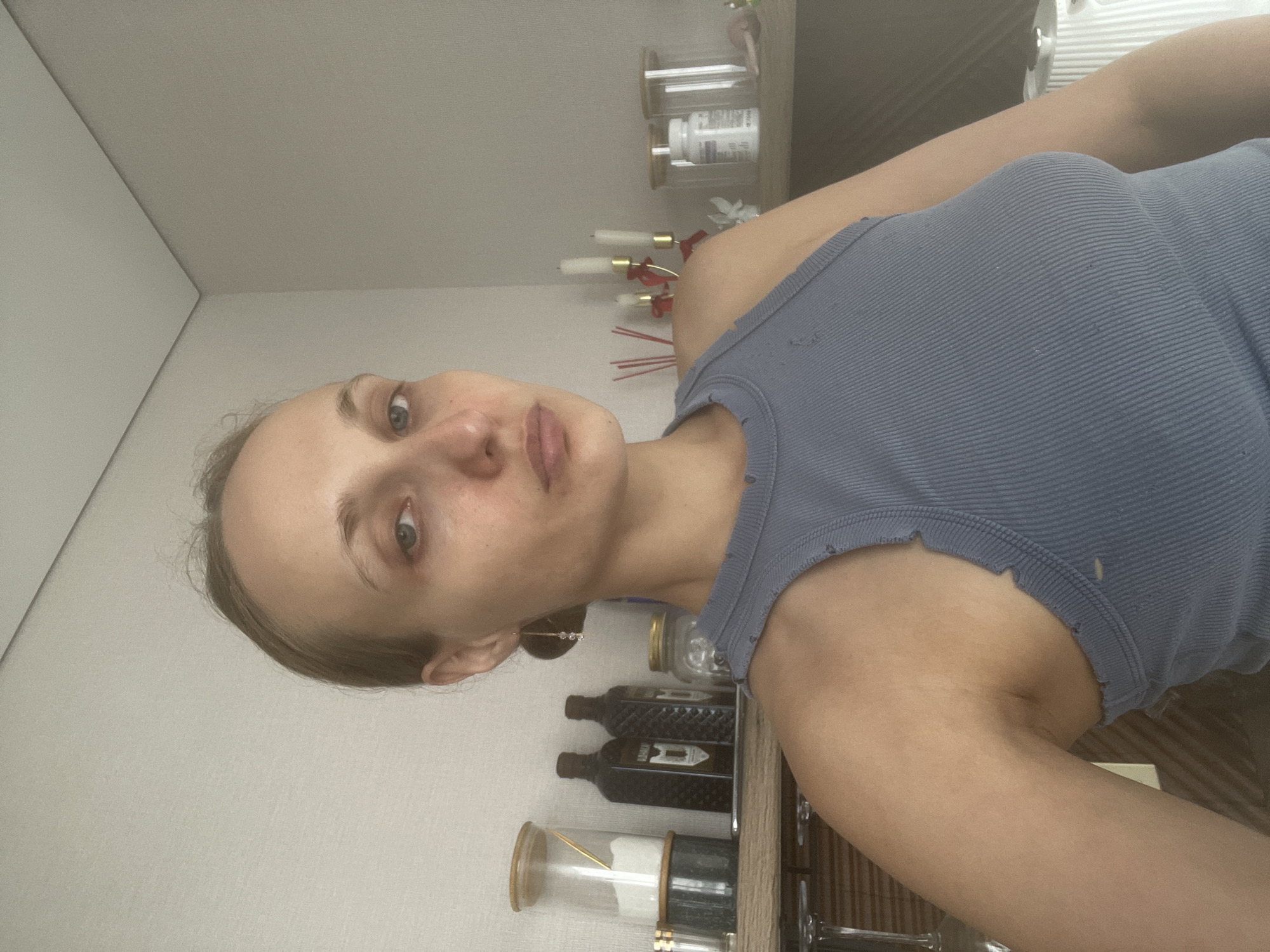 A medium shot photograph captures a woman from the chest up, seated indoors. She has fair skin, light brown hair pulled back, and is looking directly at the camera with a neutral expression. She is wearing a ribbed blue tank top with distressed detailing around the neckline and armholes. Her left arm is resting on her lap, while her right shoulder is visible. Behind her, a wooden shelf holds various objects, including glass jars with wooden lids, dark-colored bottles, and a white pill bottle. To the right of the shelf, three white pillar candles in a golden candelabra are visible. The background consists of a plain, light-colored wall with visible seams where panels meet. The overall lighting is soft and diffused, suggesting an indoor environment. The image is not a photograph of any location in the city of Подшивалово, Russia.