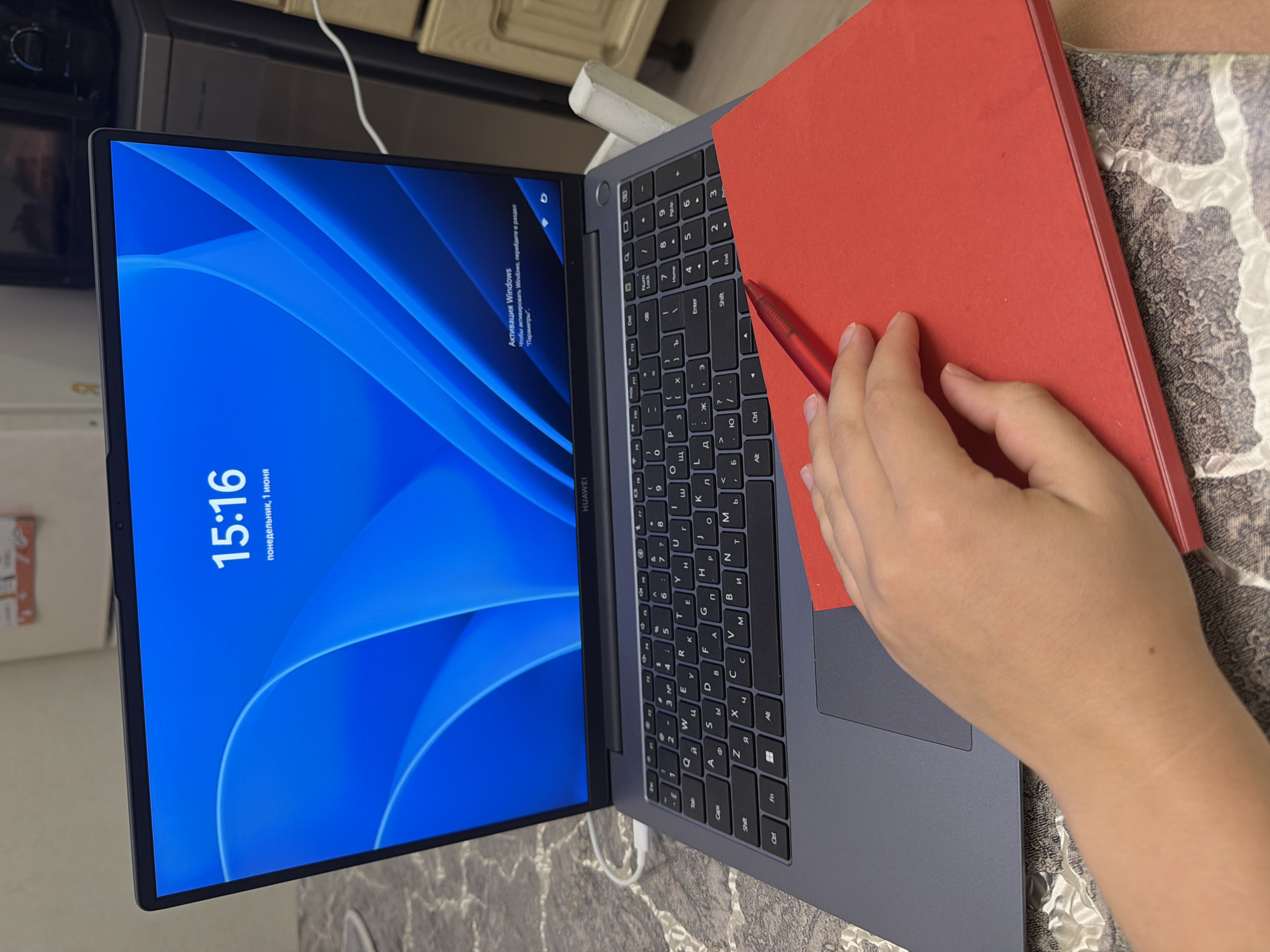 A laptop, with a red book partially covering the keyboard, sits on a patterned surface. A hand holds a red pen near the book. The laptop screen shows the time "15:16" and the date, Monday, June 1st. The laptop displays a blue graphic as a background and also displays a Windows activation message.