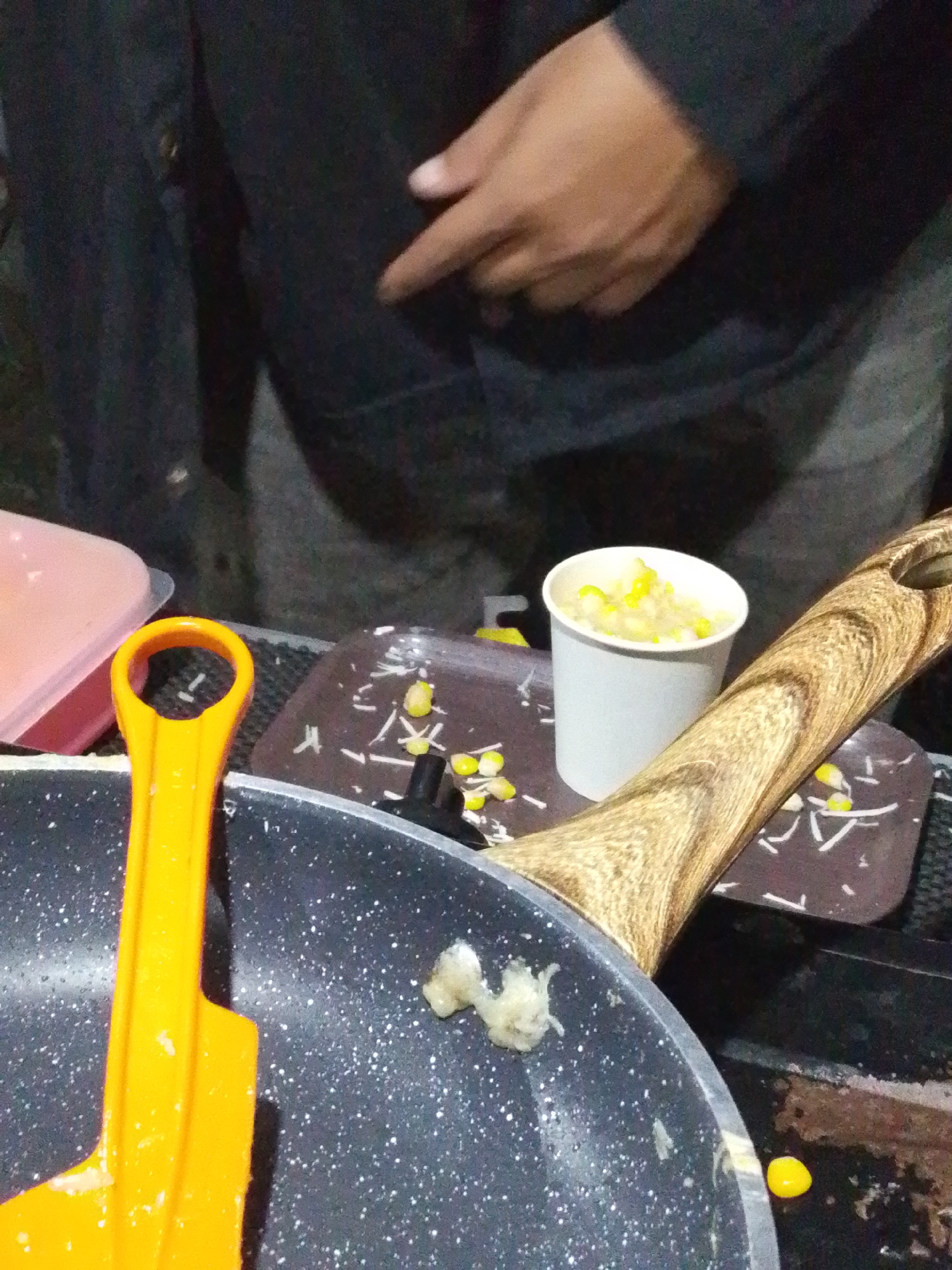 A close-up, overhead shot captures a scene likely in Lawatan, Indonesia, focused on food preparation. A dark-colored pan with white speckles occupies the foreground, with an orange spatula resting on its rim. Beside the pan, a brown tray holds scattered kernels of corn and shredded white material, possibly cheese. To the right of the tray, a white paper cup is filled with corn. A person's hand, with visible fingernails and skin tone, is seen in the upper right, seemingly reaching towards the food or a nearby object. The background is dark and out of focus, suggesting an indoor or dimly lit outdoor setting. The overall impression is one of casual food service or preparation, possibly at a street stall or outdoor event.