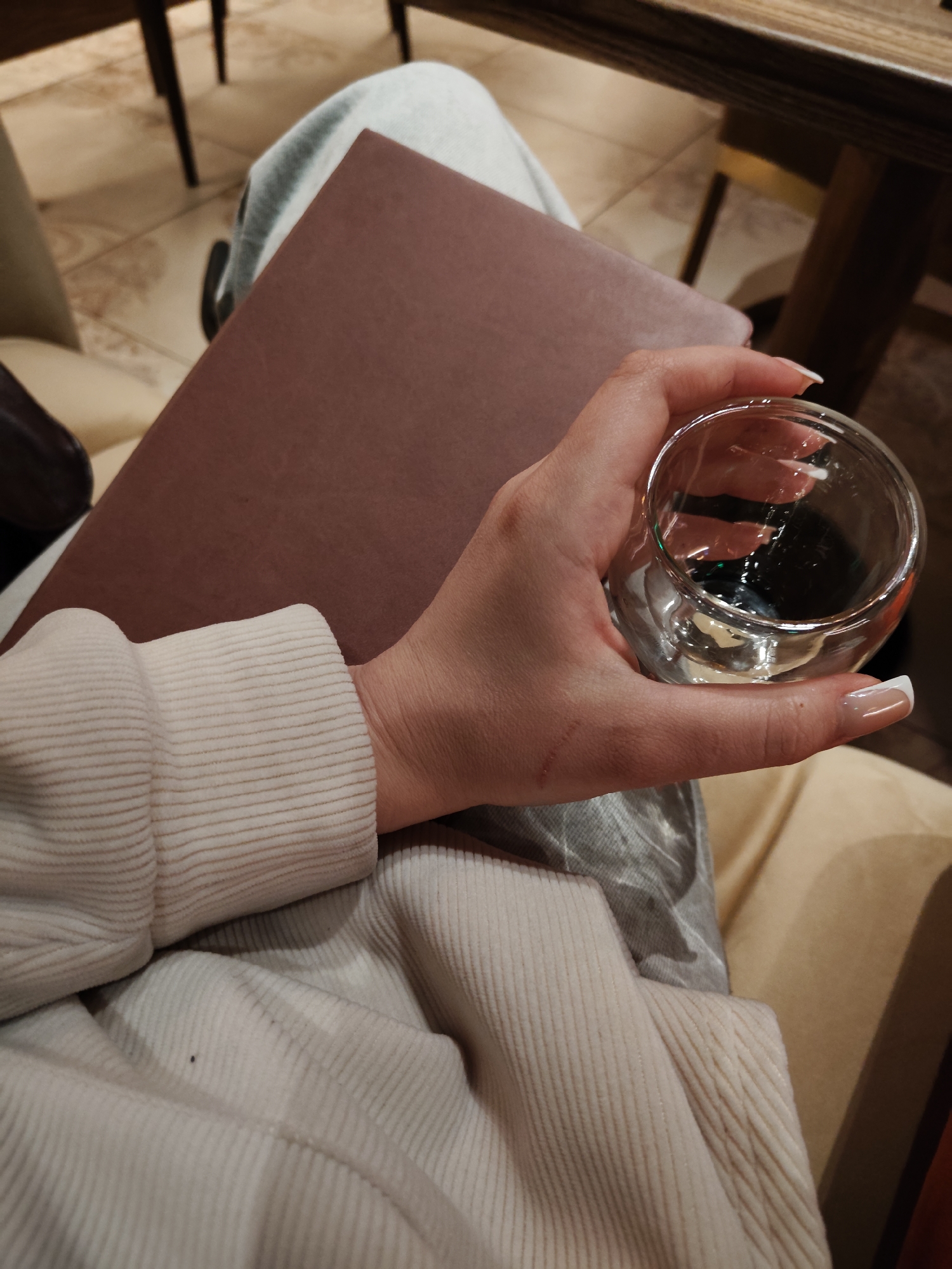 The image presents a person holding a clear glass in their right hand, with a brown, rectangular object resting on their lap. The glass contains a dark liquid and is held by a hand with manicured nails. The person is wearing a light-colored corduroy sweater. The brown object appears to be a menu or a similar document. The background suggests an indoor setting, possibly a restaurant or cafe. Furniture is visible, including a chair or bench with beige upholstery and a dark-colored table leg. The environment is dimly lit. The location is Unknown, Unknown.
