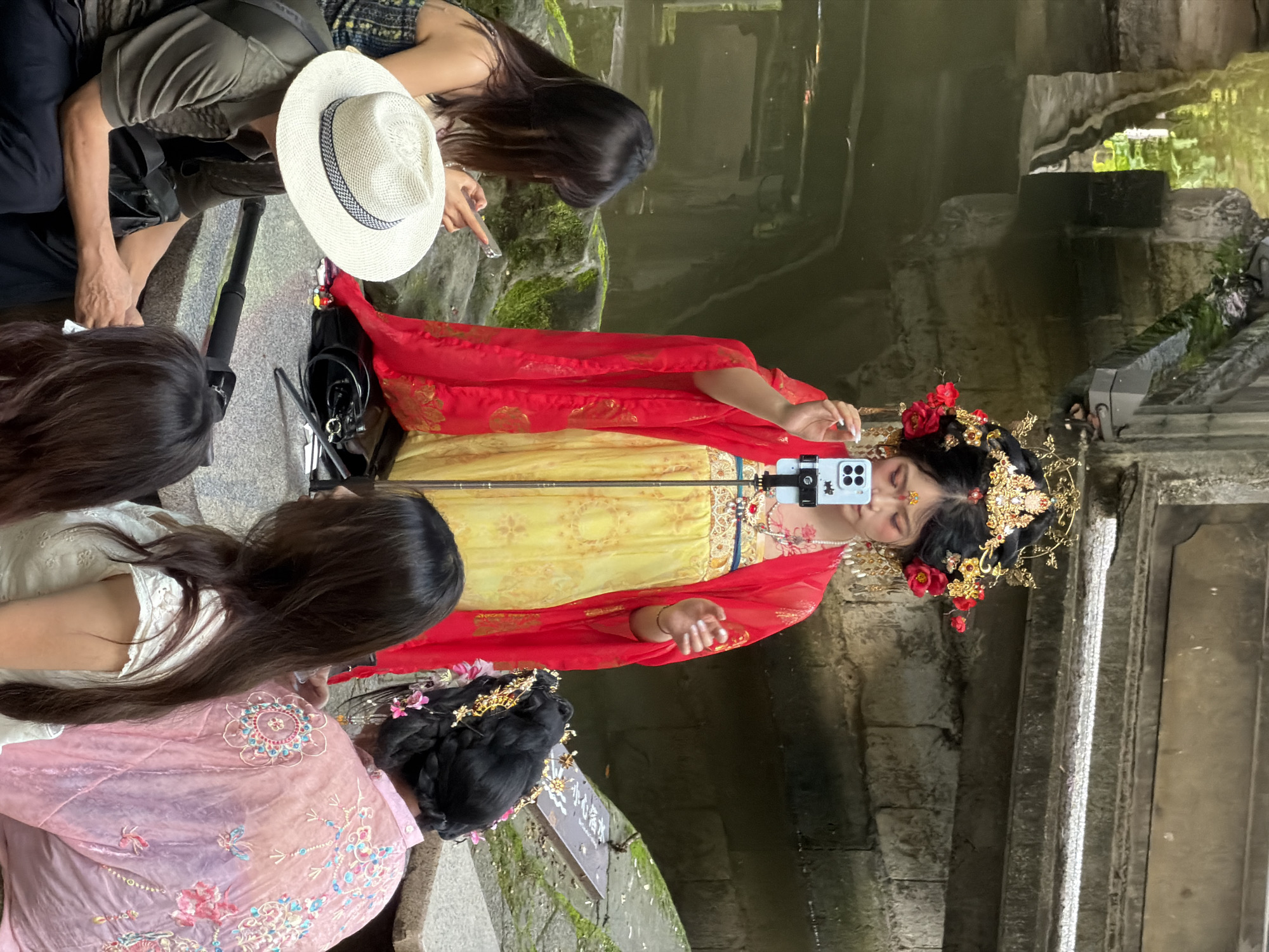 In Yuzhong District, China, a woman dressed in traditional Hanfu, adorned with an elaborate gold headpiece and red flowers, stands by a water feature. She is holding a smartphone on a selfie stick, seemingly recording herself. Around her, several individuals are present. To her left, a woman in a patterned dress looks at her phone. In front of her, a man wearing a wide-brimmed hat and a green shirt is seated, with a camera stabilizer in front of him. To the right of the woman in Hanfu, another person is seen from behind, wearing a pink embroidered garment and a traditional hairstyle with flowers. The environment appears to be a garden or park setting, with stone structures and lush greenery visible, reflecting in the water.