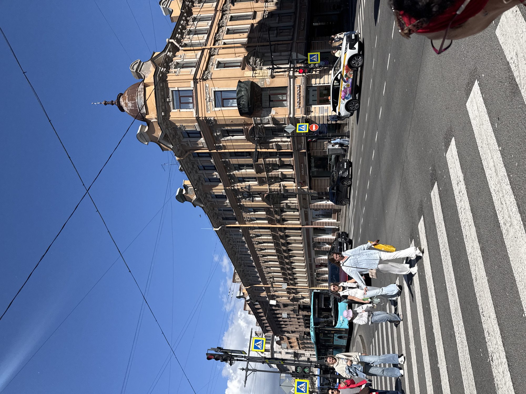This media file shows a street scene in an urban environment. The focal point is a group of people crossing a pedestrian street. The building in the background is tall and ornate, with many windows and decorative architectural details. A white car with colorful decals is parked on the right side of the street, and a blue bus is visible on the left. The sky is clear and blue, suggesting a sunny day. The people are dressed casually, and some appear to be smiling, indicating a pleasant atmosphere. There are traffic lights and street signs visible, with some text on them that is not clearly legible due to the image quality and orientation. The presence of a pedestrian crossing indicates a busy urban area with pedestrian traffic. The overall impression is that of a lively city street on a bright day.
