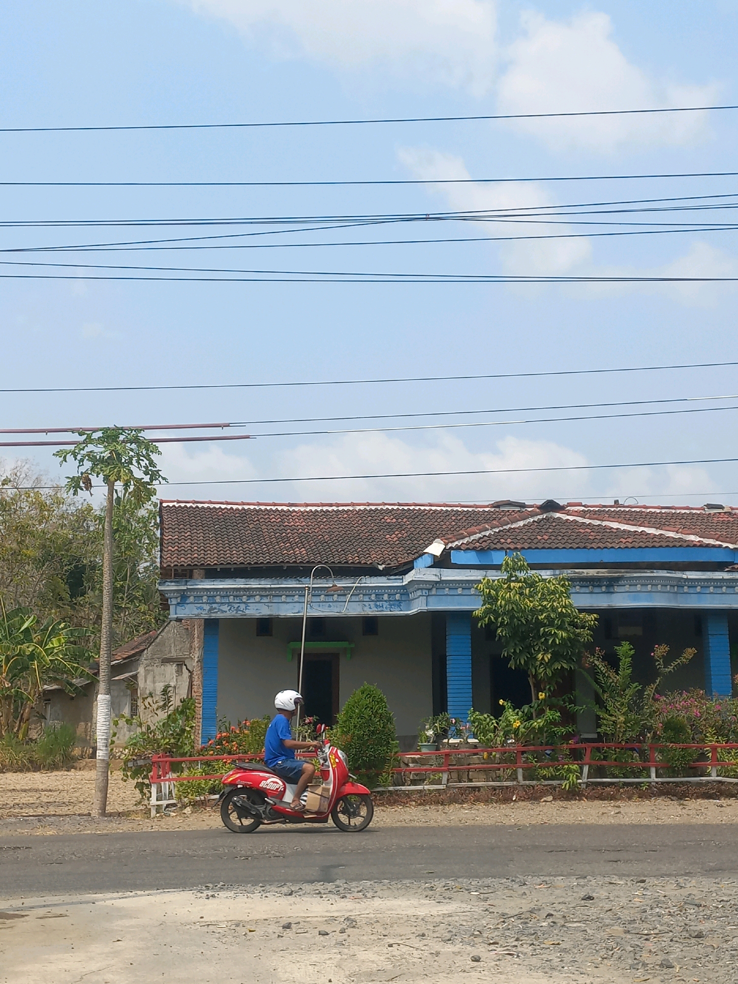 A person wearing a white helmet and a blue t-shirt rides a red scooter on a paved road. The scooter has the word "SCOOPY" written on its side. Behind the road, there is a house with a red tiled roof and blue trim. A fence made of red wooden posts and planks runs in front of the house, separating it from the road. Various plants and trees are visible in front of and around the house. In the background, there are power lines stretching across a light blue sky with scattered clouds. The area appears to be in a rural or suburban setting, possibly in Baturetno, Indonesia, given the context.