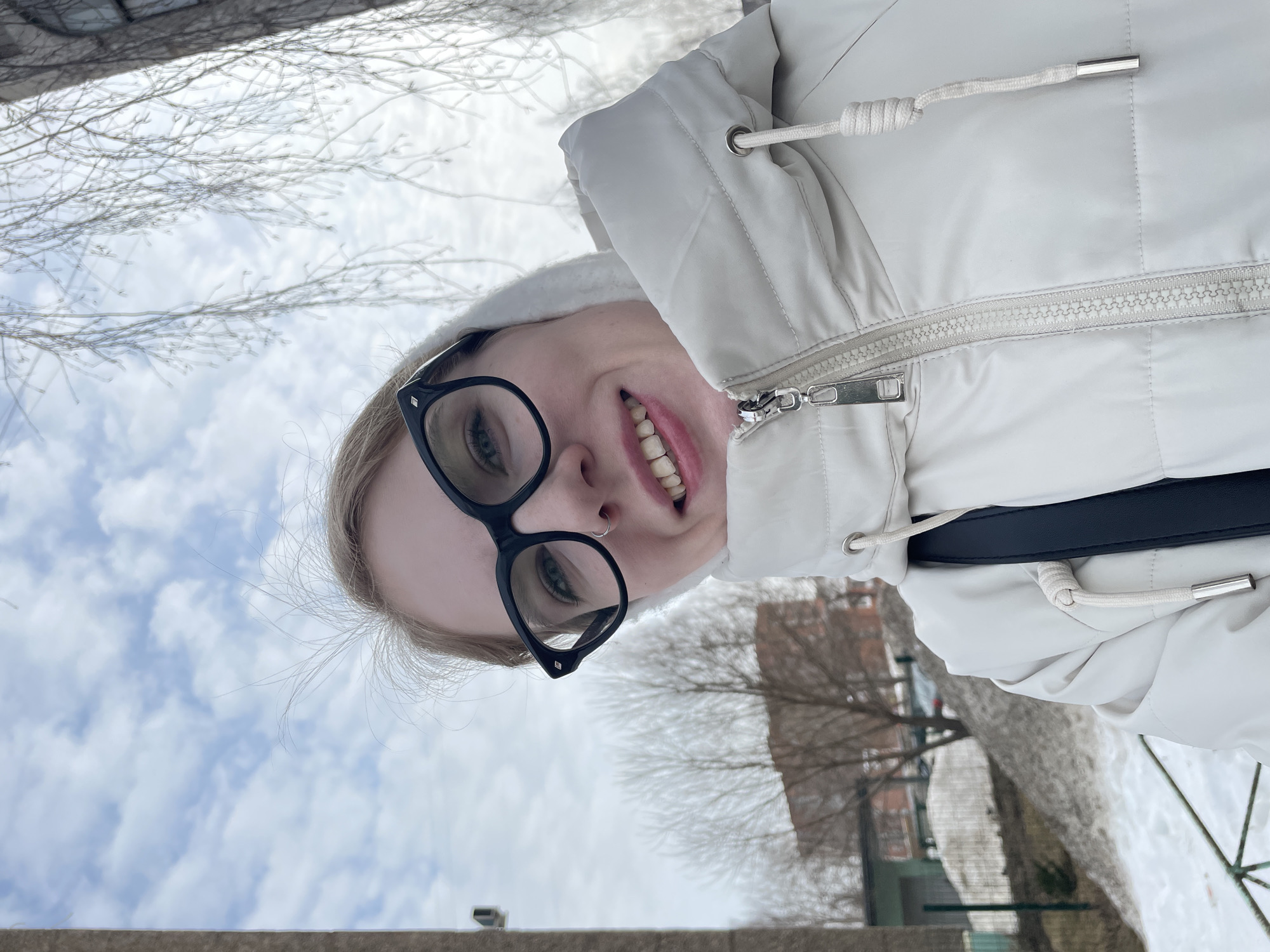 An individual with blonde hair, wearing black-framed glasses and a small silver nose ring in the right nostril, is depicted in a close-up, upward-angled self-portrait. The individual displays a smile, revealing the upper dentition. They are dressed in a cream-colored quilted winter jacket with a visible zipper and drawstrings; a dark shoulder strap is visible over the left shoulder. The background features a partially cloudy sky. To the right, bare tree branches exhibiting a crystalline coating consistent with rime ice or frost extend downwards. To the left, a vertical, light-gray concrete structure is partially visible. The mid-ground comprises snow accumulations, several deciduous trees without foliage, and a multi-story building with a reddish-brown facade in the distance. A low green fence and a compact green structure are present on the left. A metal railing is partially visible in the lower-left foreground. The scene exhibits characteristics of a cold environment, including snow cover and bare trees, some with crystalline deposits, indicative of late winter or early spring. While the general urban environment is consistent with a city like Izhevsk, Russia, the specific location within Izhevsk cannot be determined from the provided visual information alone.
