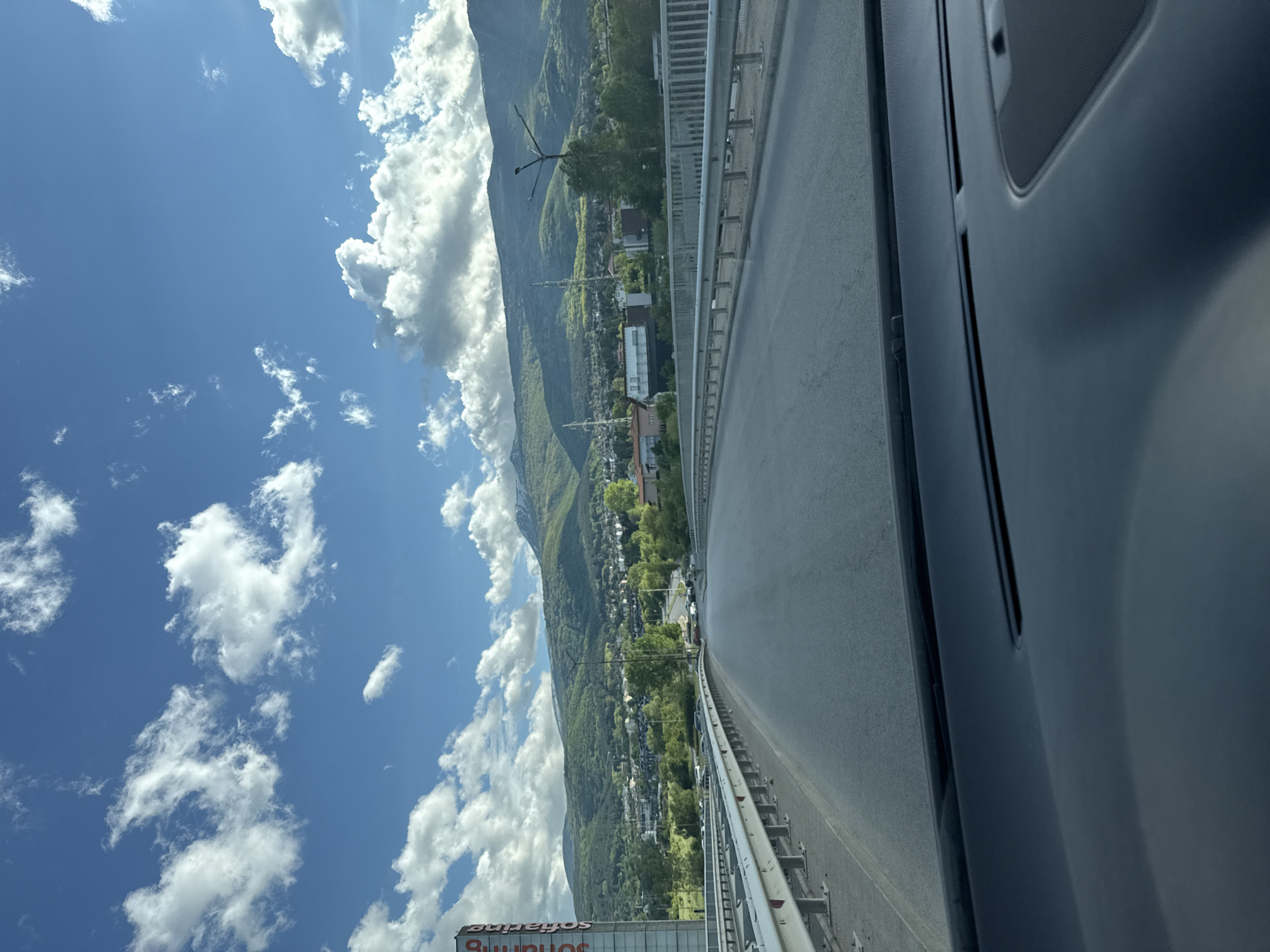 The image shows a view from a car driving on a bridge, with a highway on the right side of the frame. A guardrail separates the highway from a landscape featuring mountains and a city in the distance. The sky is blue with scattered white clouds. The car's interior is visible on the right side of the image, suggesting the photo was taken from inside the vehicle. There is a sign visible. Based on the sign in the image showing "Johannes", the location is likely somewhere in Bulgaria, as the only place that fits the description from the information provided is Sofia.