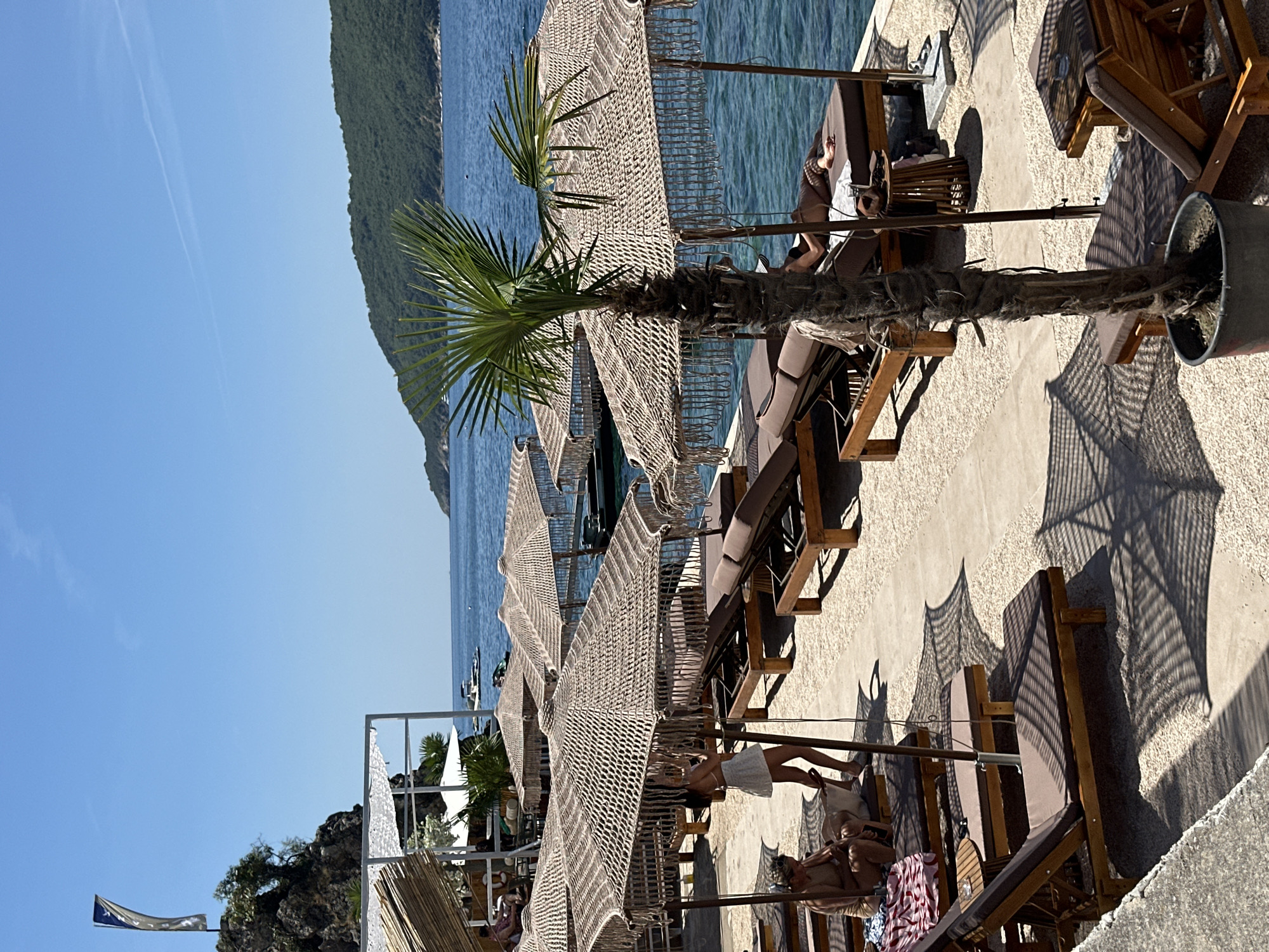 This image depicts a sunny day at a beachfront resort in Budva, Montenegro. The scene features rows of empty and occupied lounge chairs with thatched umbrellas, set on a gravelly beach next to the clear blue sea. A few people are visible relaxing on the loungers. A small palm tree stands in the foreground, casting a shadow on the beach. In the background, lush green hills meet the horizon, and a few boats are dotted on the water. A flag is visible on a rocky outcrop to the left. The overall atmosphere is one of leisure and relaxation.