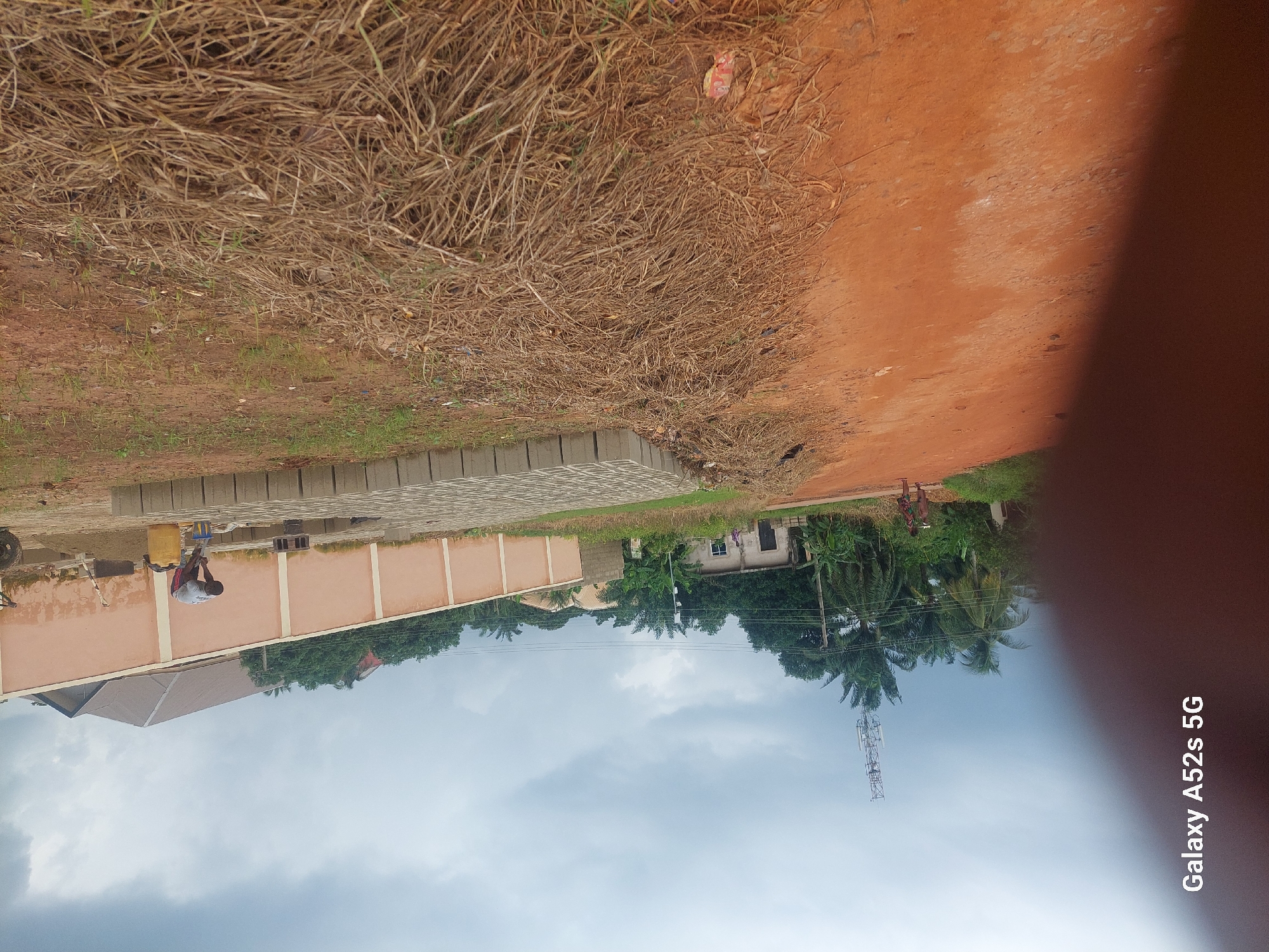 The image depicts an outdoor scene, likely in Umudioka, Nigeria. A person is visible in the mid-ground, engaged in an activity near a building under construction. The building has a pale orange facade and a dark grey roof. Adjacent to the building, a stack of grey concrete blocks is arranged in a line, suggesting construction work is in progress. Dry, brown vegetation and reddish-brown soil dominate the foreground and right side of the image. In the background, a tall telecommunications tower stands against an overcast sky. Trees, including palm trees, and smaller buildings are scattered throughout the scene. A red dirt road is present on the right. The image also shows a watermark at the bottom left corner, indicating it was captured with a "Galaxy A52s 5G" phone.