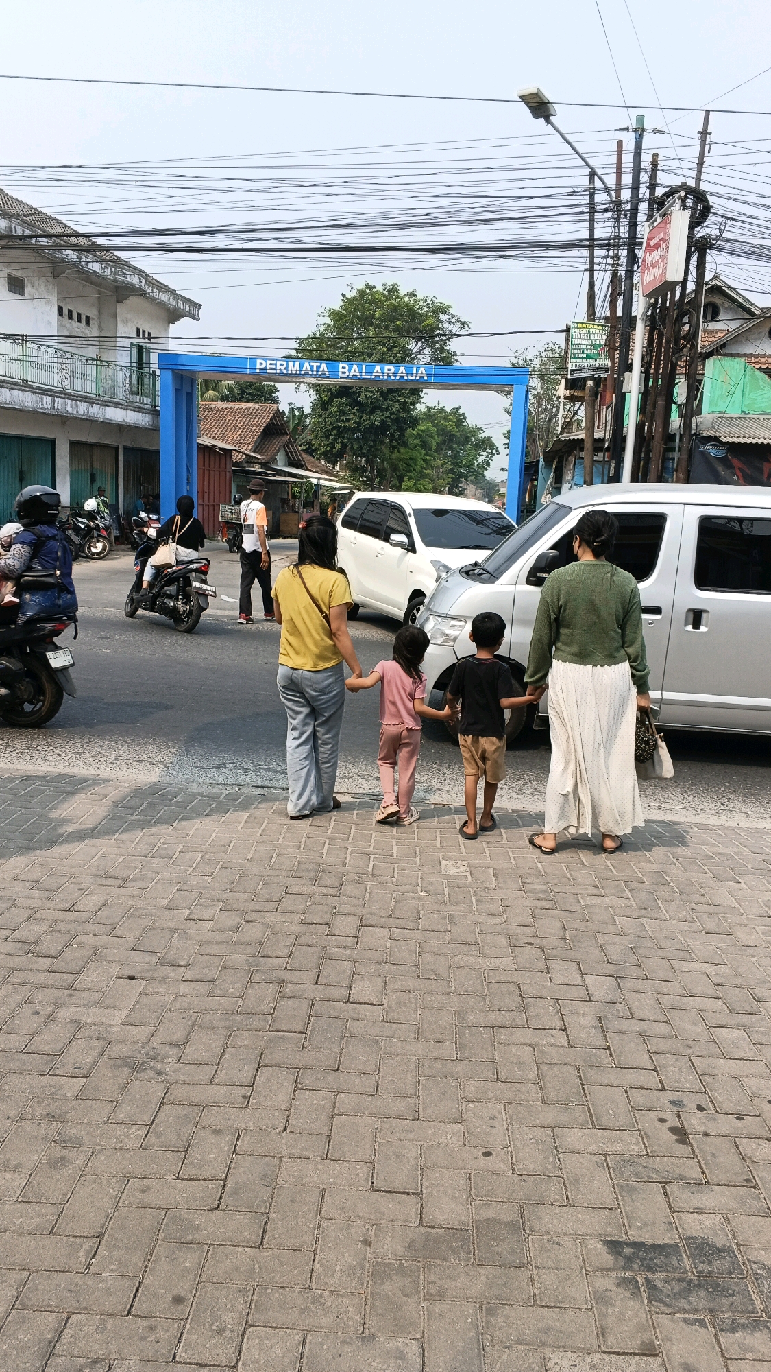 The image depicts a street scene in Balaraja, Indonesia, with a prominent blue archway bearing the text "PERMATA BALARAJA". In the foreground, two women and two children are holding hands and walking across a brick-paved sidewalk. The woman on the left wears a yellow t-shirt and grey pants, while the woman on the right is dressed in a green long-sleeved shirt and a long white skirt. The children, a boy and a girl, are dressed in casual clothing. Behind them, traffic flows on the asphalt road, with several motorcycles and two white vehicles visible. The environment appears to be urban, with buildings and utility poles lining the street. The sky is overcast, and the lighting suggests it is daytime.