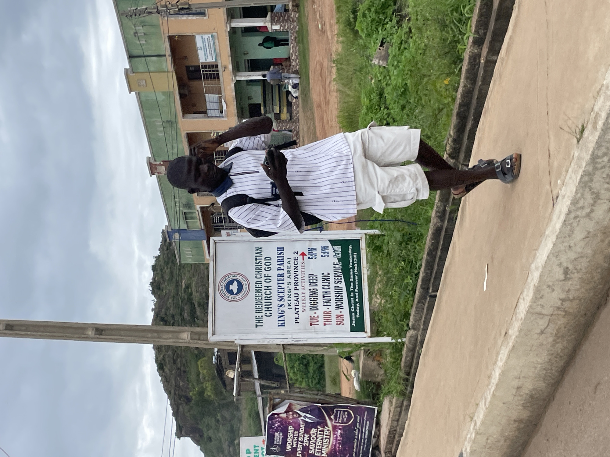 A dark-skinned man, wearing a white striped shirt with blue stripes, white shorts, and dark sandals, is walking on a paved walkway in front of a building. He is holding a dark-colored smartphone to his right ear with his right hand, and his left hand is also near the phone. He is carrying a black backpack. To his left is a white sign with black text listing church activities for "THE REDEEMED CHRISTIAN CHURCH OF GOD KING'S SCEPTRER PARISH". The sign also includes the church's logo, a dove, and a Bible verse. Behind the man are two buildings, one painted orange and yellow, and another section painted green. The sky is overcast and cloudy. In the background, there is a rocky, vegetated hill. The man is walking towards the right of the frame. This scene is located in Gura Top, Nigeria.