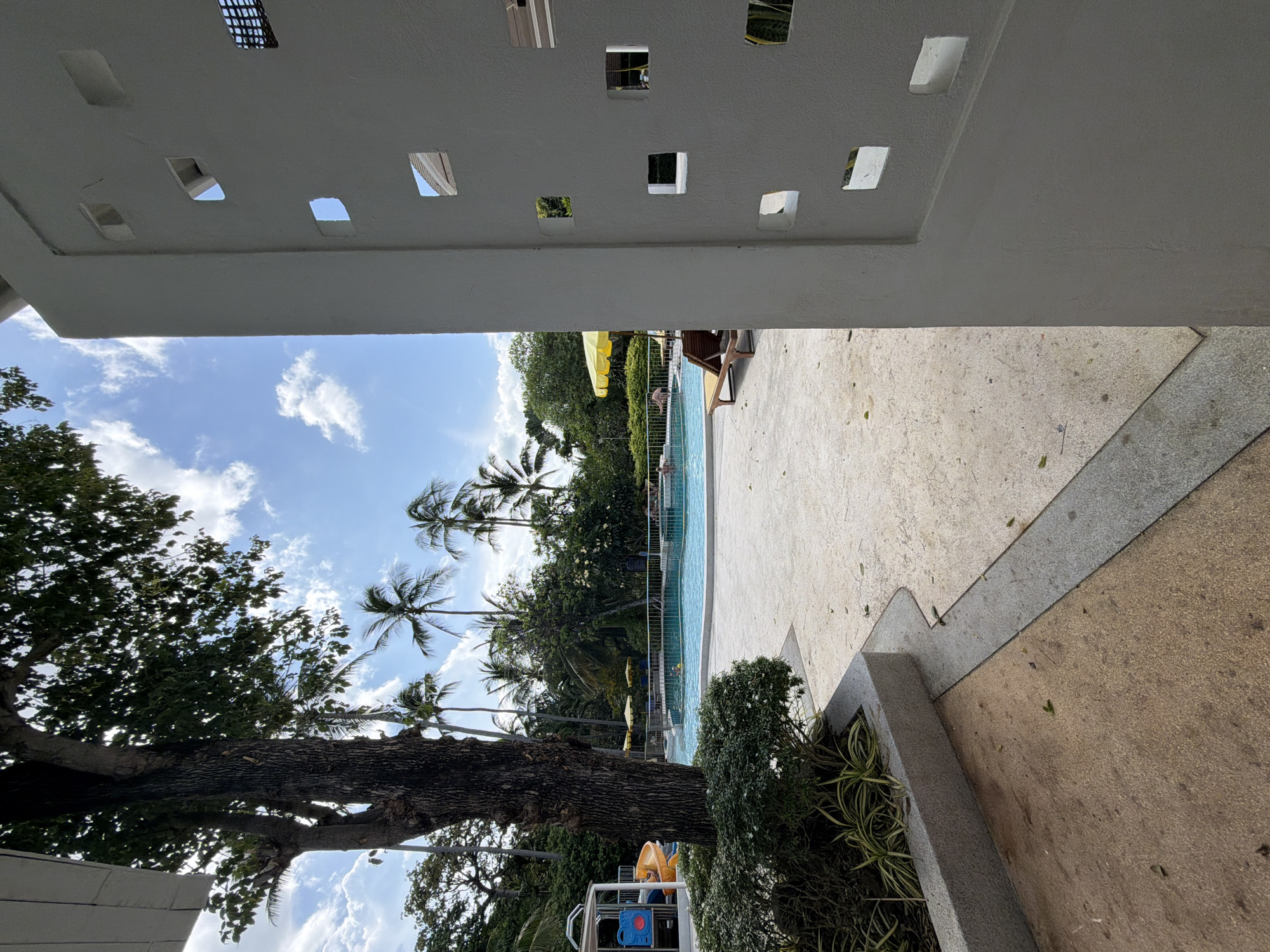 The image depicts a swimming pool area at a resort in Koh Samui, Thailand, on a sunny day with scattered clouds. In the foreground, a white wall with square cutouts partially obstructs the view. To the left, a large tree with dark bark stands prominently, with lush green bushes at its base. Beyond the tree, a bright blue swimming pool is visible, with several people swimming and relaxing. Yellow umbrellas provide shade over lounge chairs by the pool. A children's play structure with a slide is also present. Palm trees and other tropical foliage surround the pool area, creating a serene and inviting atmosphere. The ground is paved with light-colored concrete. The overall impression is that of a well-maintained resort amenity.