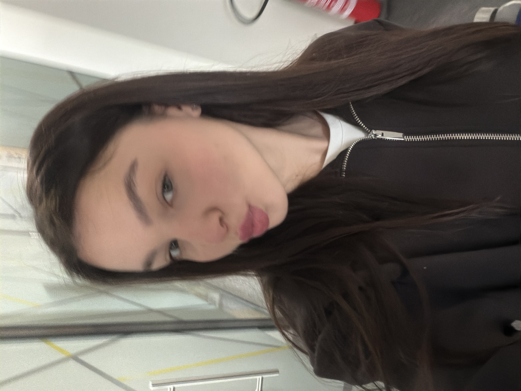 A medium shot shows a young woman with dark hair wearing a black jacket with a white shirt underneath. The background suggests a bright, interior space, possibly a waiting area with a window and some metal fixtures. There is no visible indication to confirm that the location is Sofia, Bulgaria.