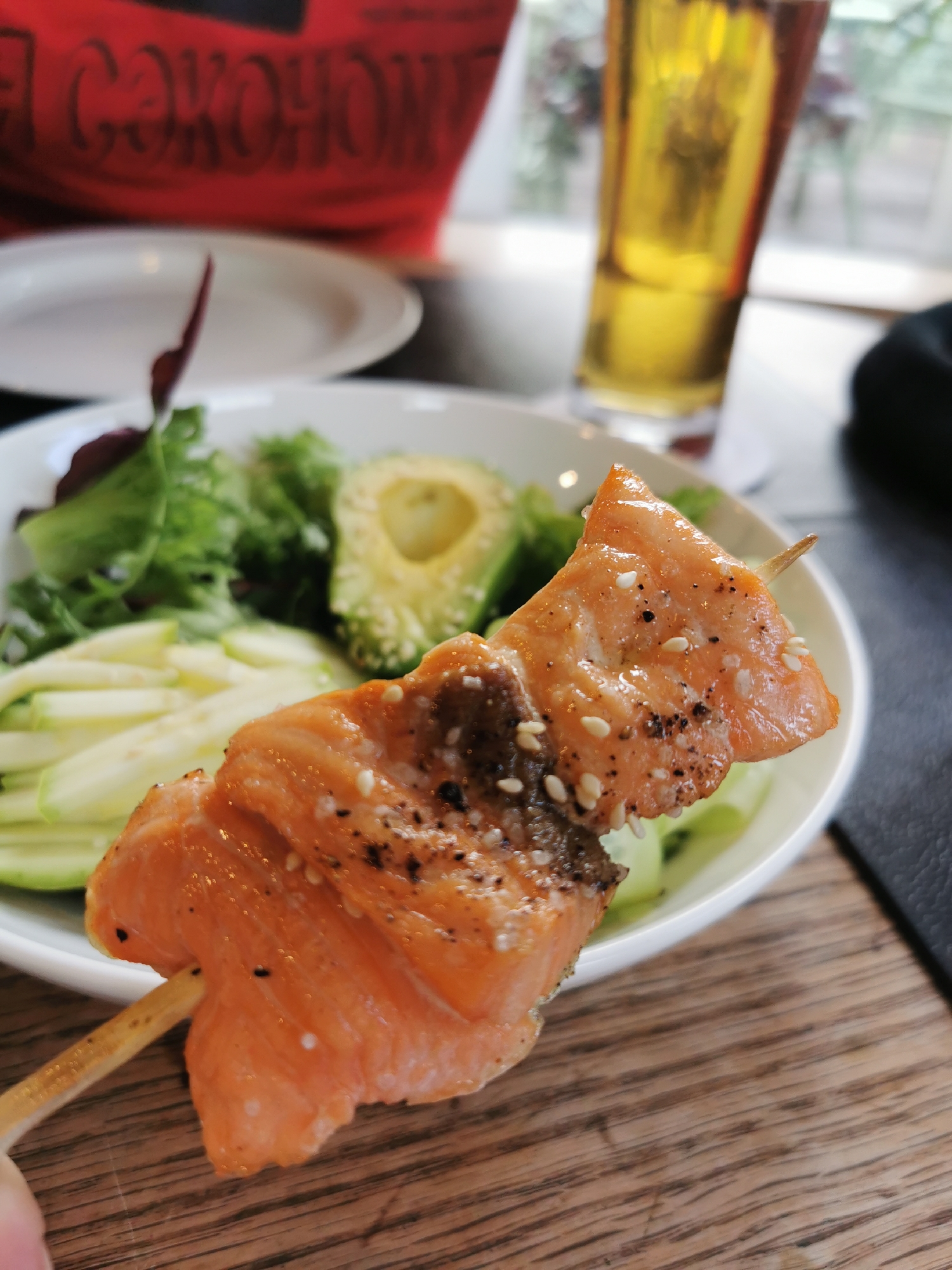 The image depicts a close-up of a salmon skewer held in someone's hand, positioned in front of a white bowl containing a salad. The salad includes shredded light green vegetables, leafy greens, and a halved avocado. Behind the bowl, out of focus, is a tall glass of beer and a person wearing a red t-shirt with writing on it. In the background, light is filtering through what appears to be a window with a blurred outdoor scene visible. The table has a wooden surface. There is also a white plate visible in the blurred background. The location is likely a restaurant or dining establishment, possibly in Moscow, Russia given the inscription on the t-shirt appears to be in the Cyrillic alphabet.
